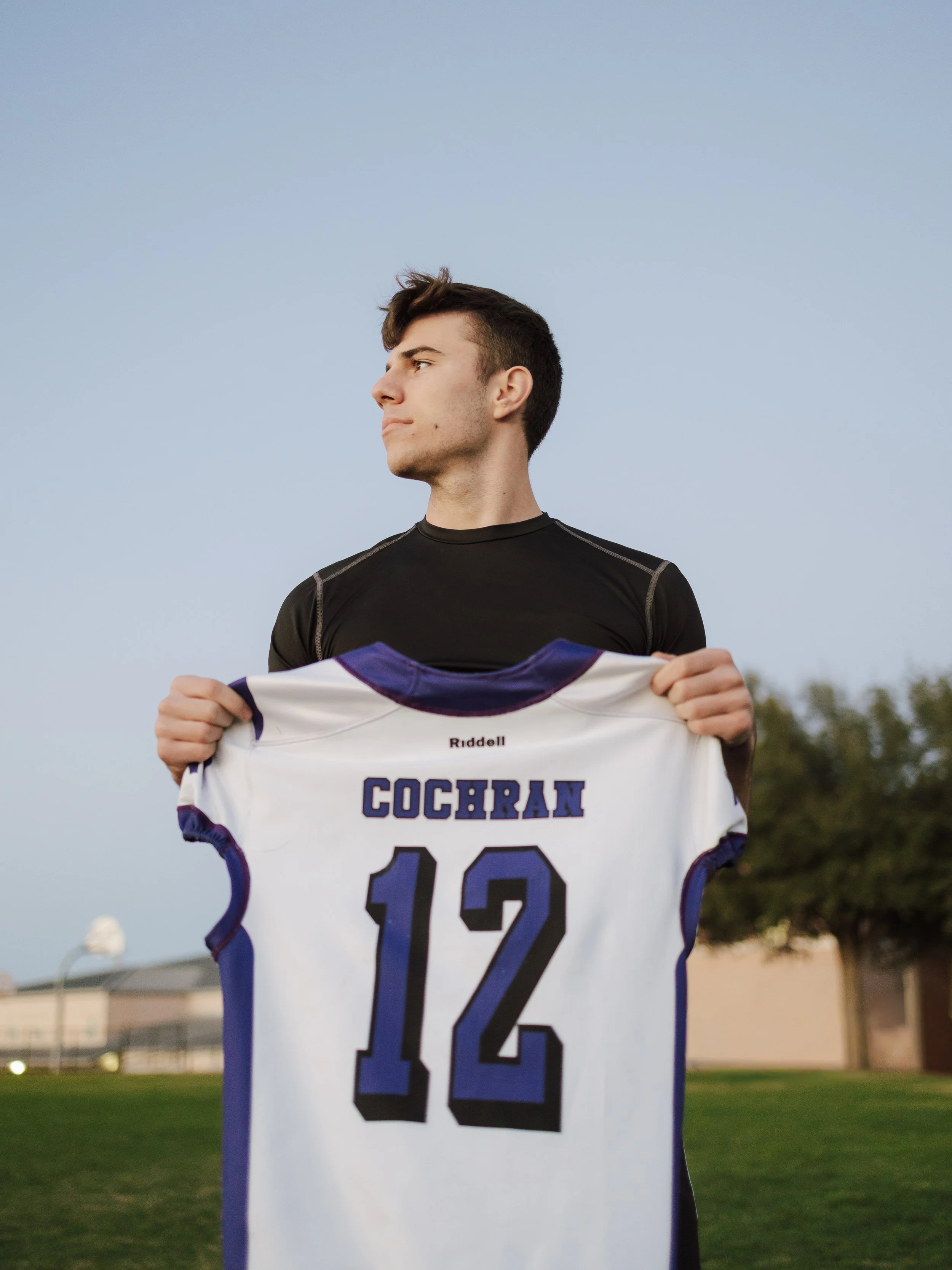 Young man holding a sports jersey with the number 12, standing outdoors during the evening.