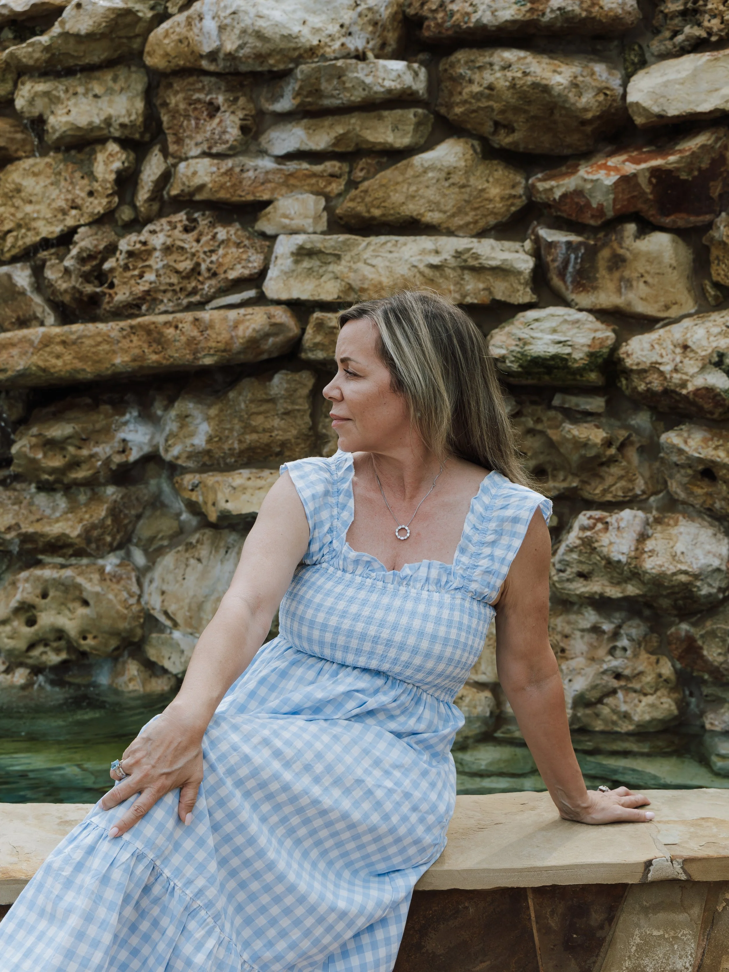 A woman in a blue and white checkered dress sitting on a stone bench near a stone wall, with her hands resting on the bench and she is looking to the side.