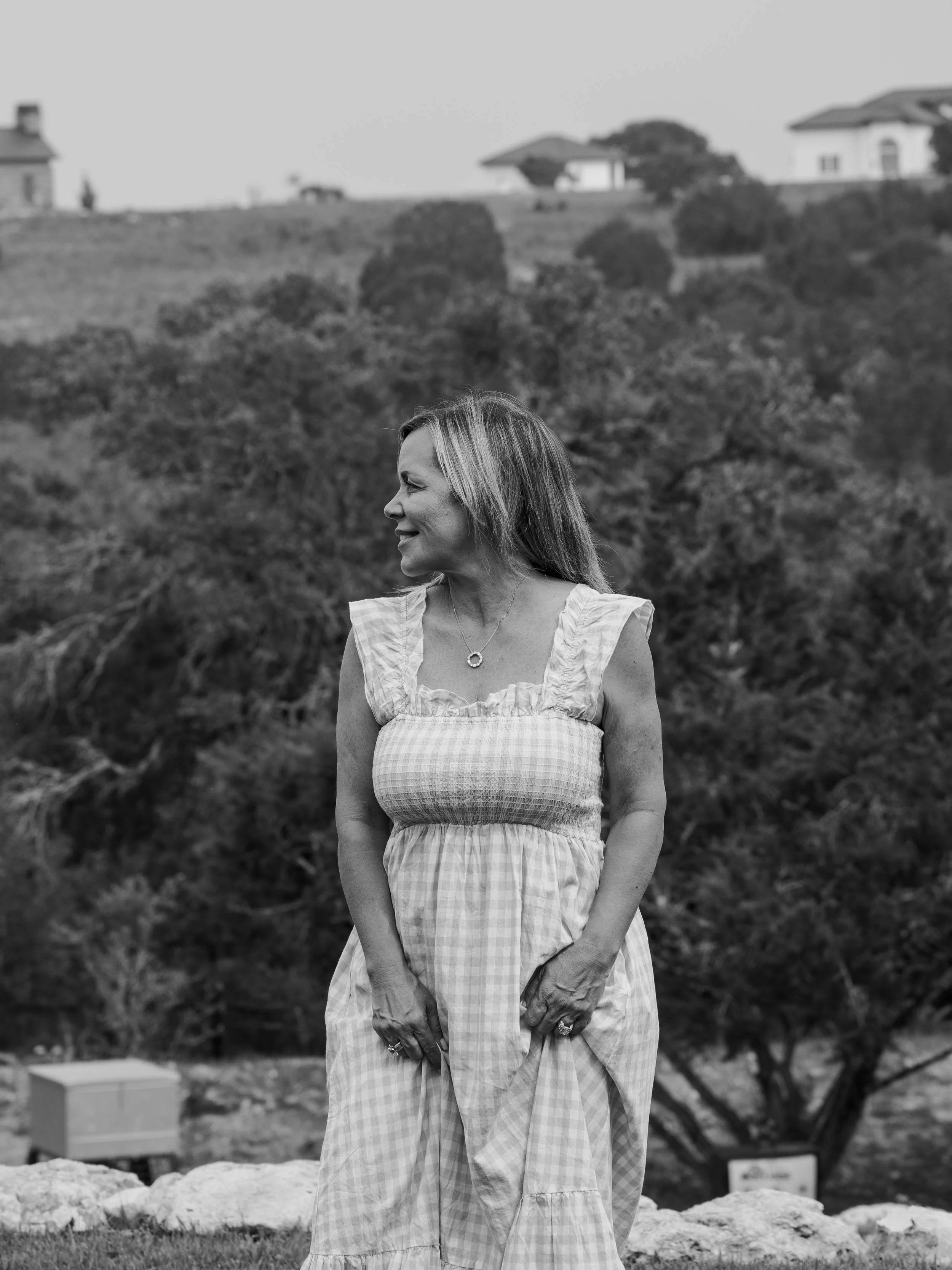 A woman with blonde hair smiling and looking to her left, outdoors with trees and houses in the background, wearing a light-colored dress with ruffles and a necklace.