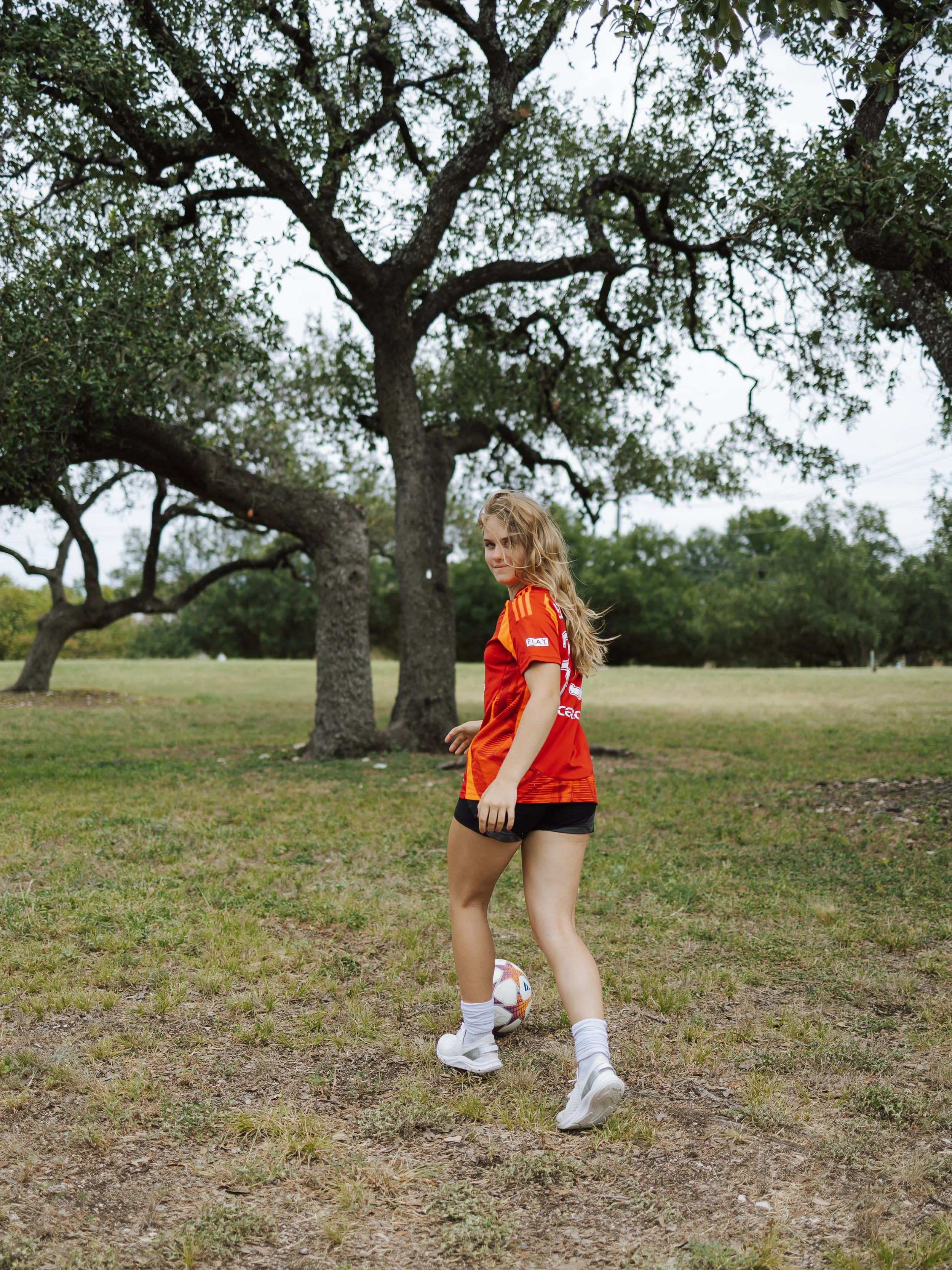 A young woman wearing a red sports jersey and black shorts standing outdoors on grass with a soccer ball near her foot, under a large tree with abundant green foliage.