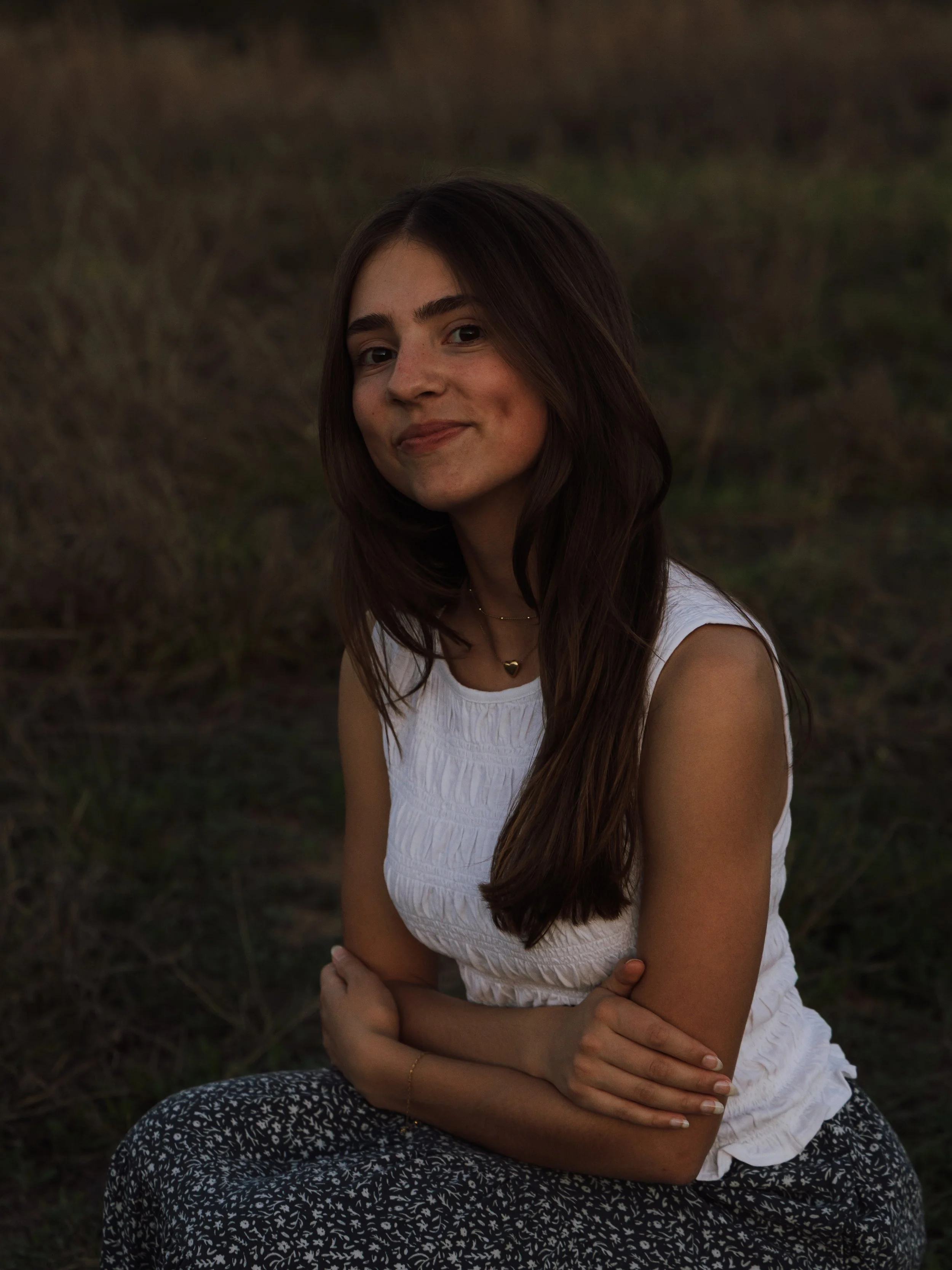 A young woman sitting outdoors on grass, wearing a white sleeveless top and patterned pants, with her arms crossed, smiling softly at the camera during what appears to be dusk or early evening.