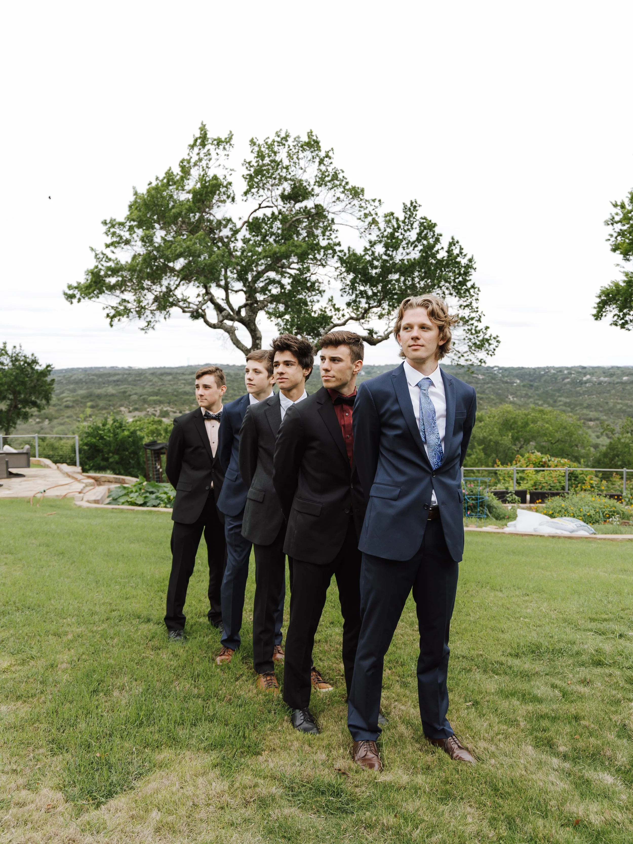 Group of young men in formal suits standing in a line outdoors with a large tree and a lawn in the background.