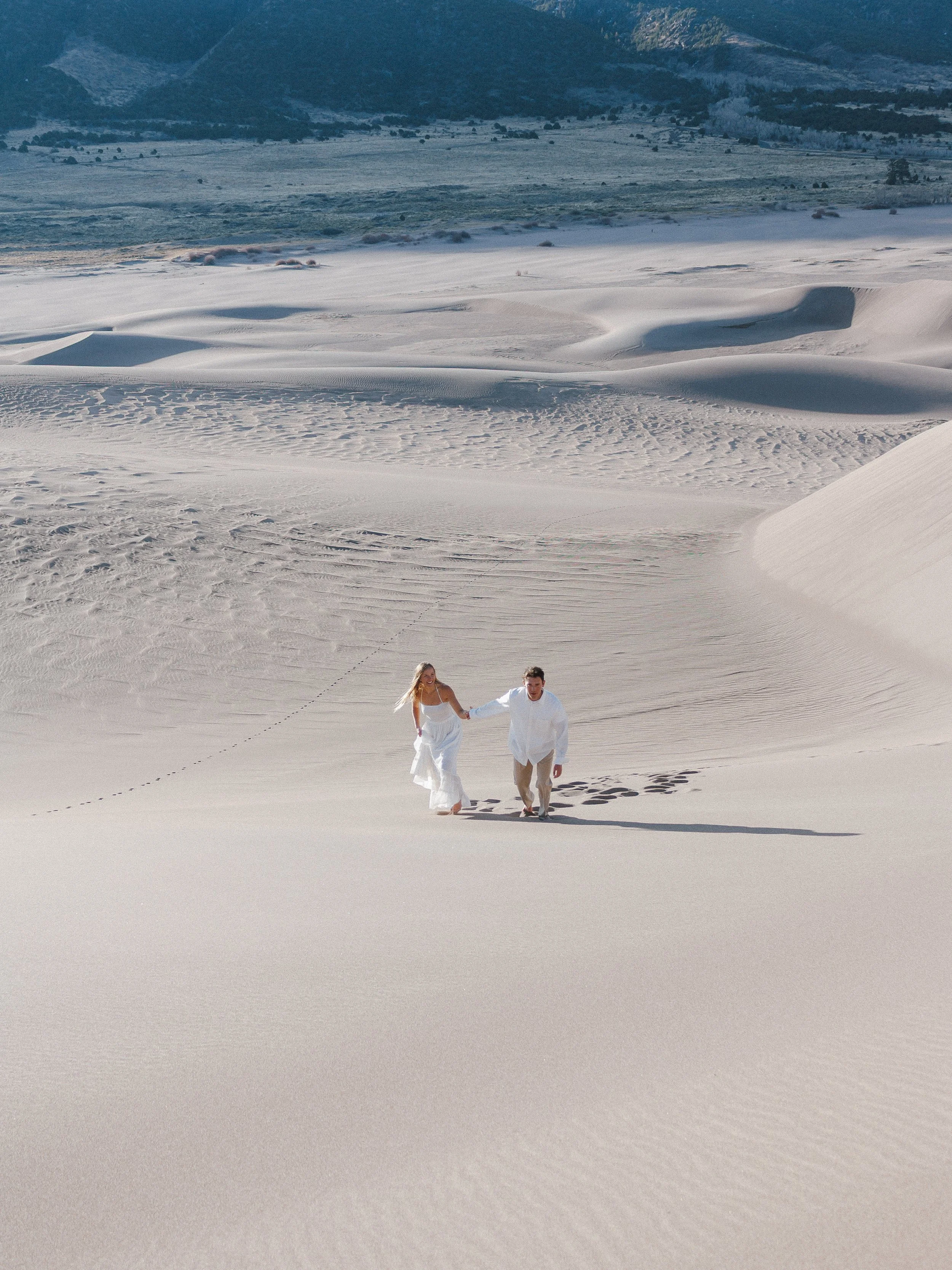 A couple holding hands while walking on white sand dunes in a desert landscape.