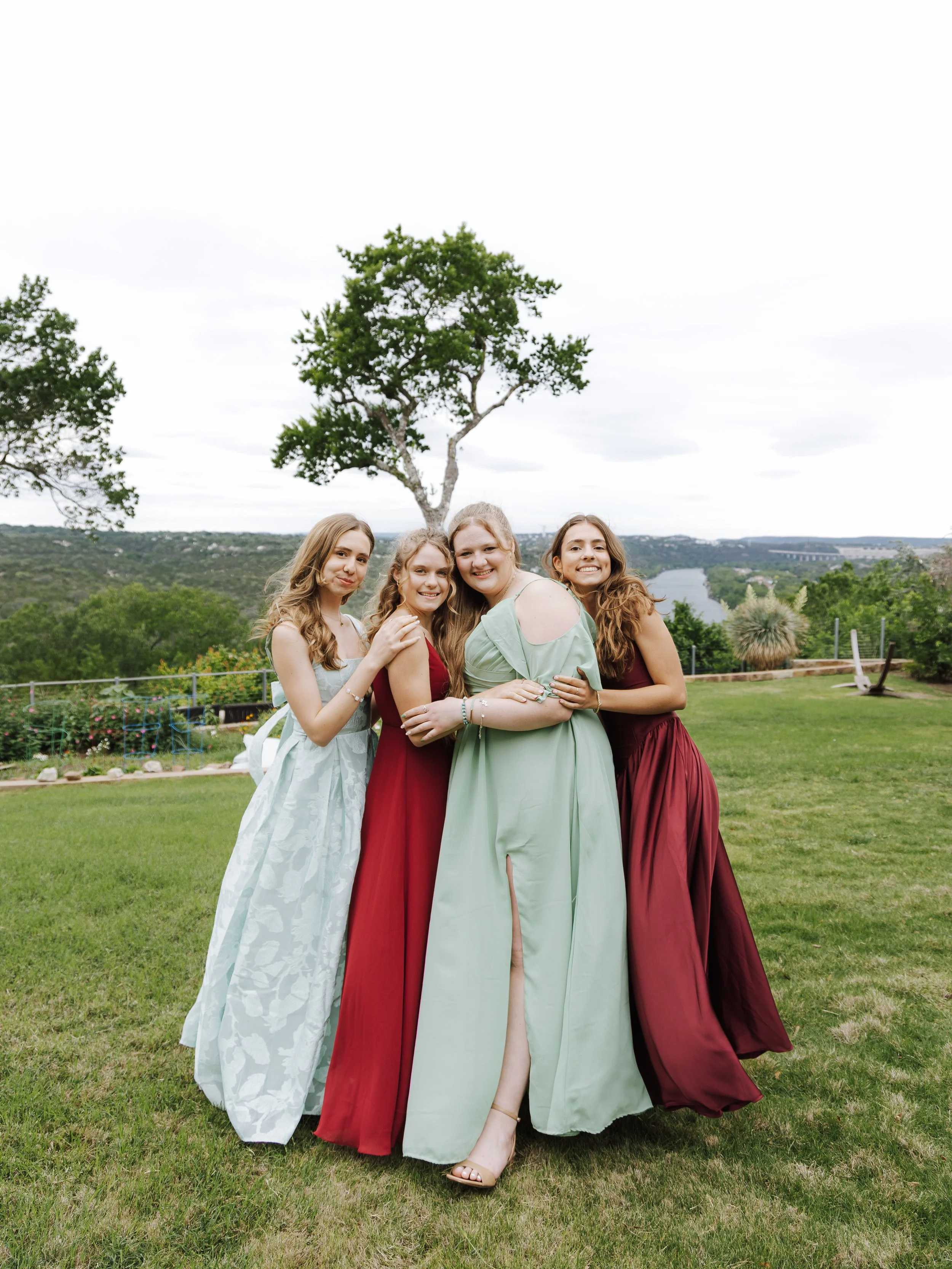 Four young women standing together outdoors on a grassy hill, wearing long gowns in pastel and burgundy colors, smiling with arms around each other, with trees and a lake in the background.