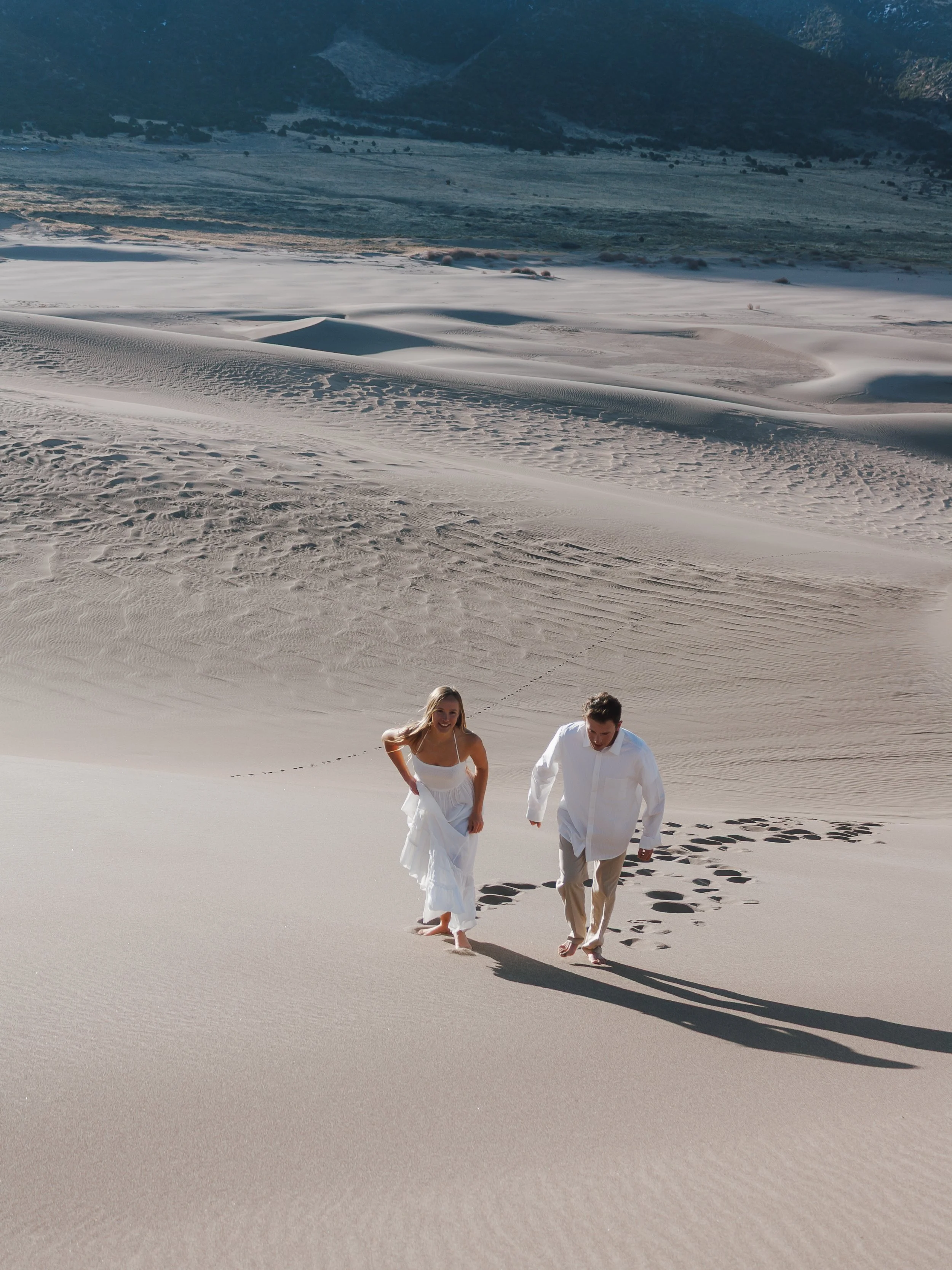 A couple dressed in white walking barefoot on sand dunes in a desert landscape with mountains in the background.