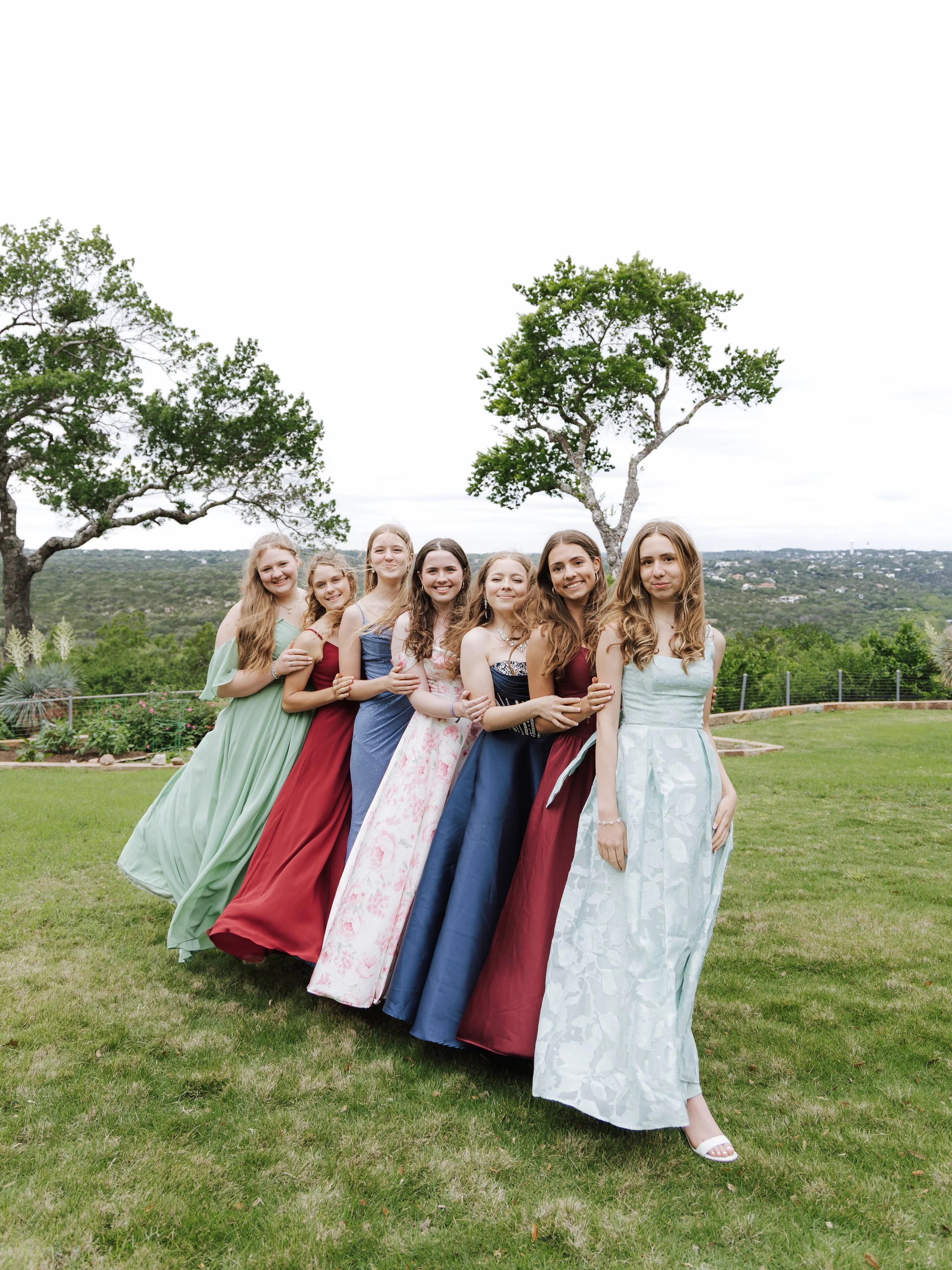 Group of young women in colorful dresses standing outdoors on a grassy hill with trees and a view of the landscape in the background.
