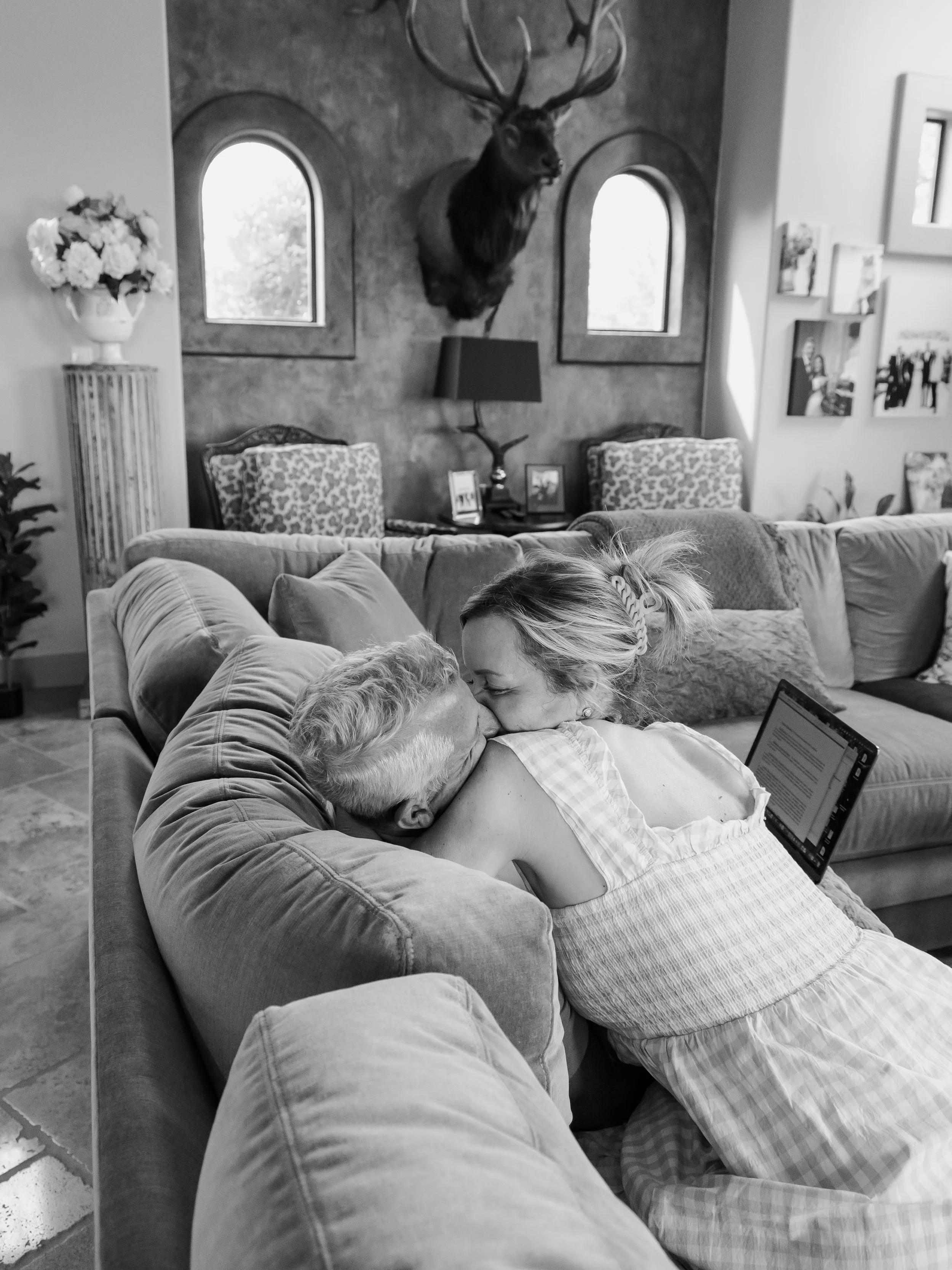 A woman and an elderly man are sitting on a sofa, sharing a kiss in a cozy living room. The woman has a tablet in her lap. There is a deer head mounted on the wall behind them, with two arched windows on either side. A floral arrangement is visible o