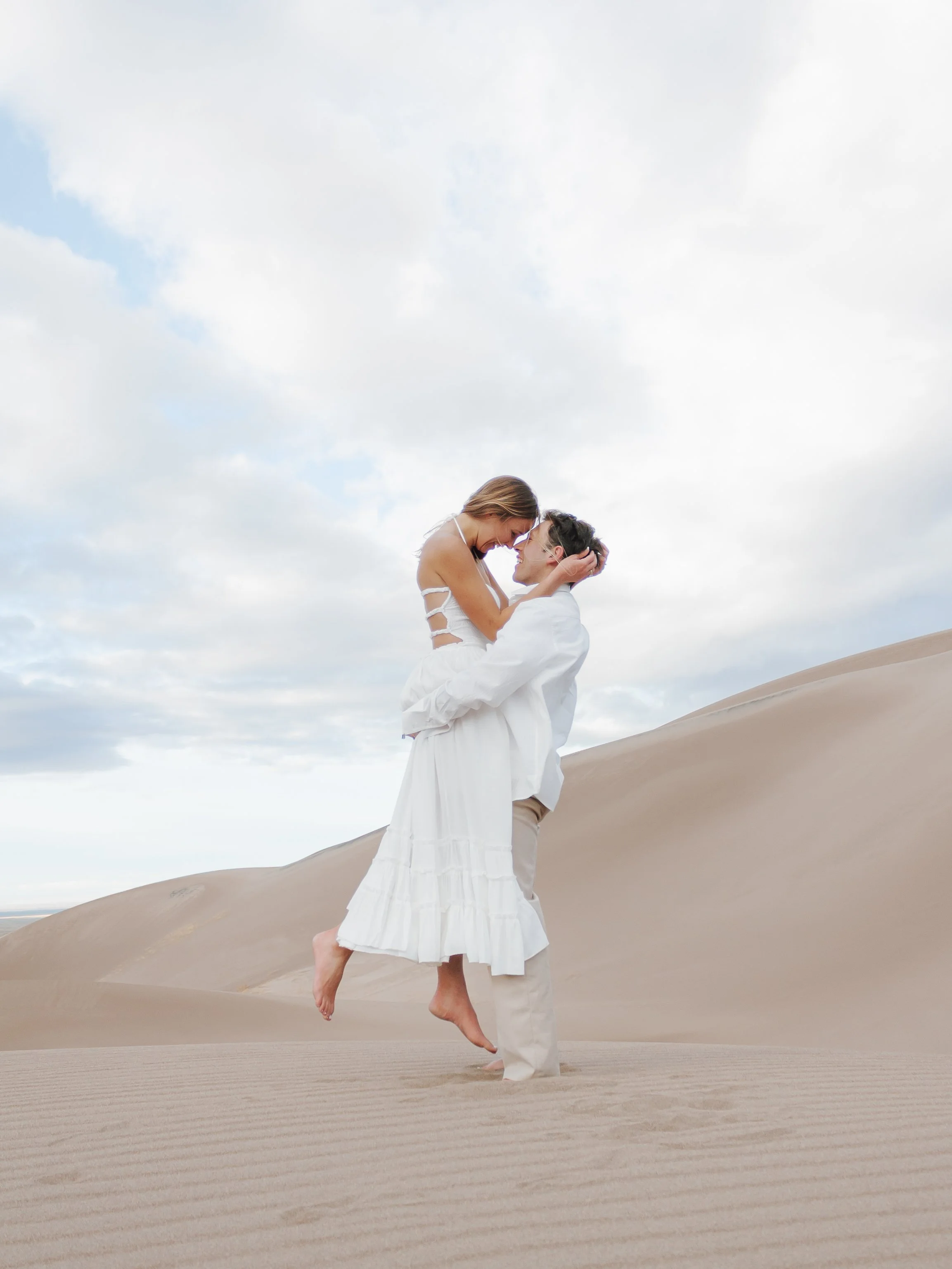 A couple in white clothing celebrates in a desert with sand dunes under a cloudy sky, with the man lifting the woman off the ground.