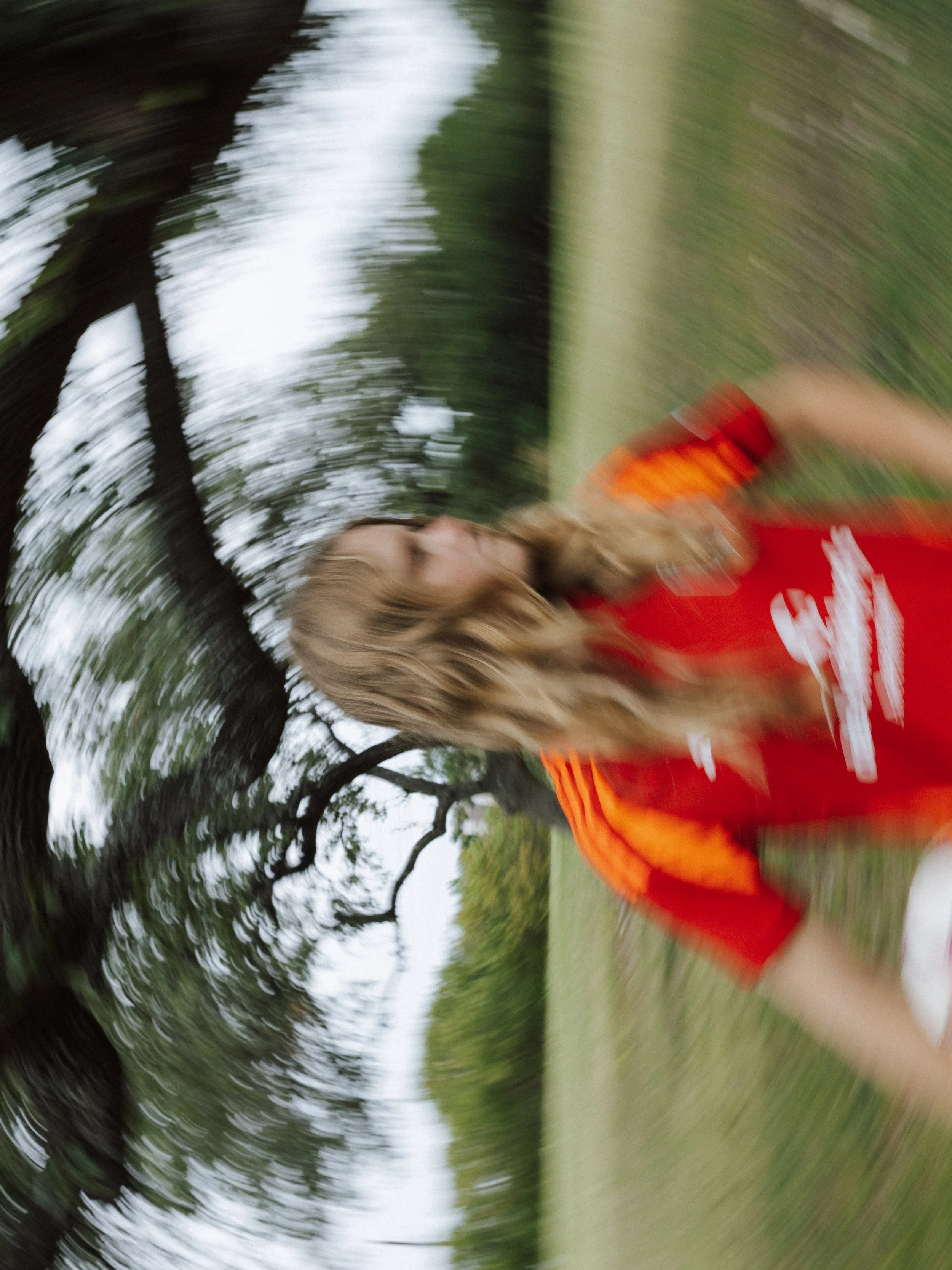 Blurred image of a blonde woman in a red and orange sports shirt running outdoors on a grassy field with trees in the background.