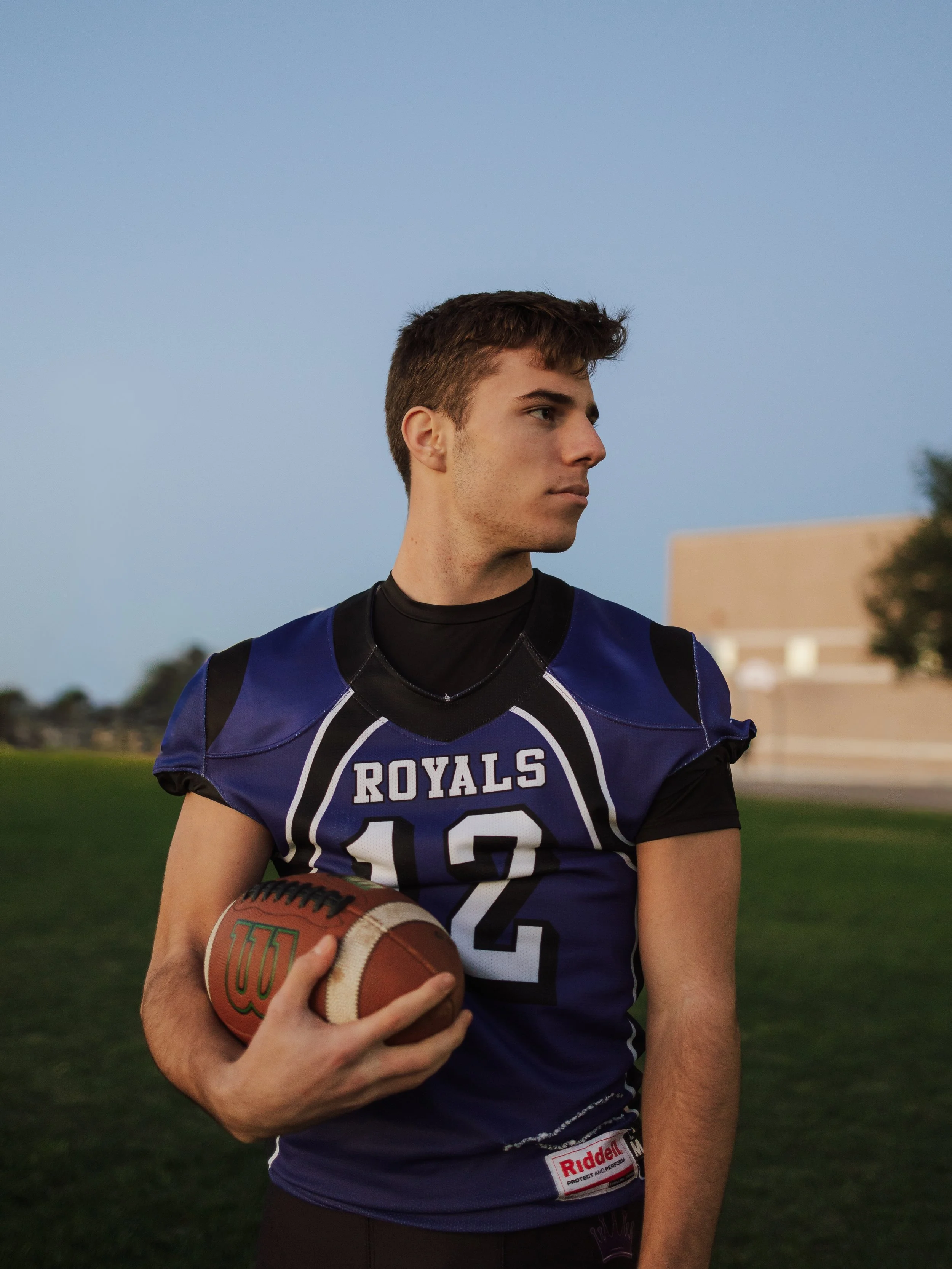 A young man stands outdoors on a sports field at dusk, holding a football, wearing a blue and black football jersey with the word "Royals" and the number "12" on it.