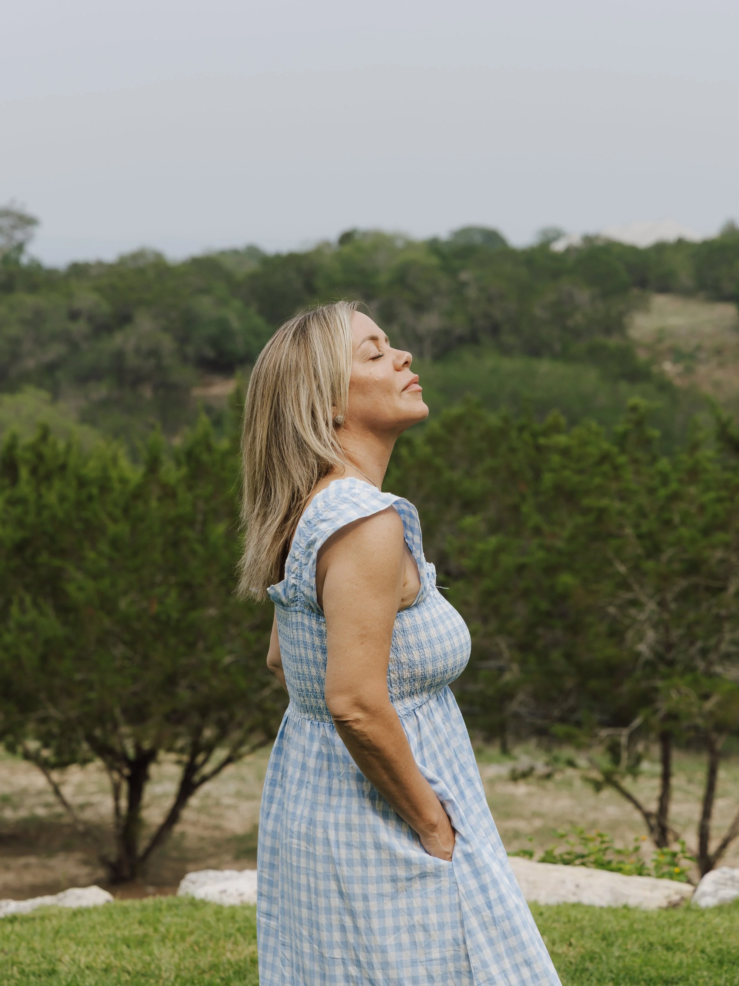 A woman with blonde hair, wearing a blue and white checkered sleeveless dress, standing outdoors with her eyes closed and face turned slightly upward, surrounded by green trees and grass on a cloudy day.
