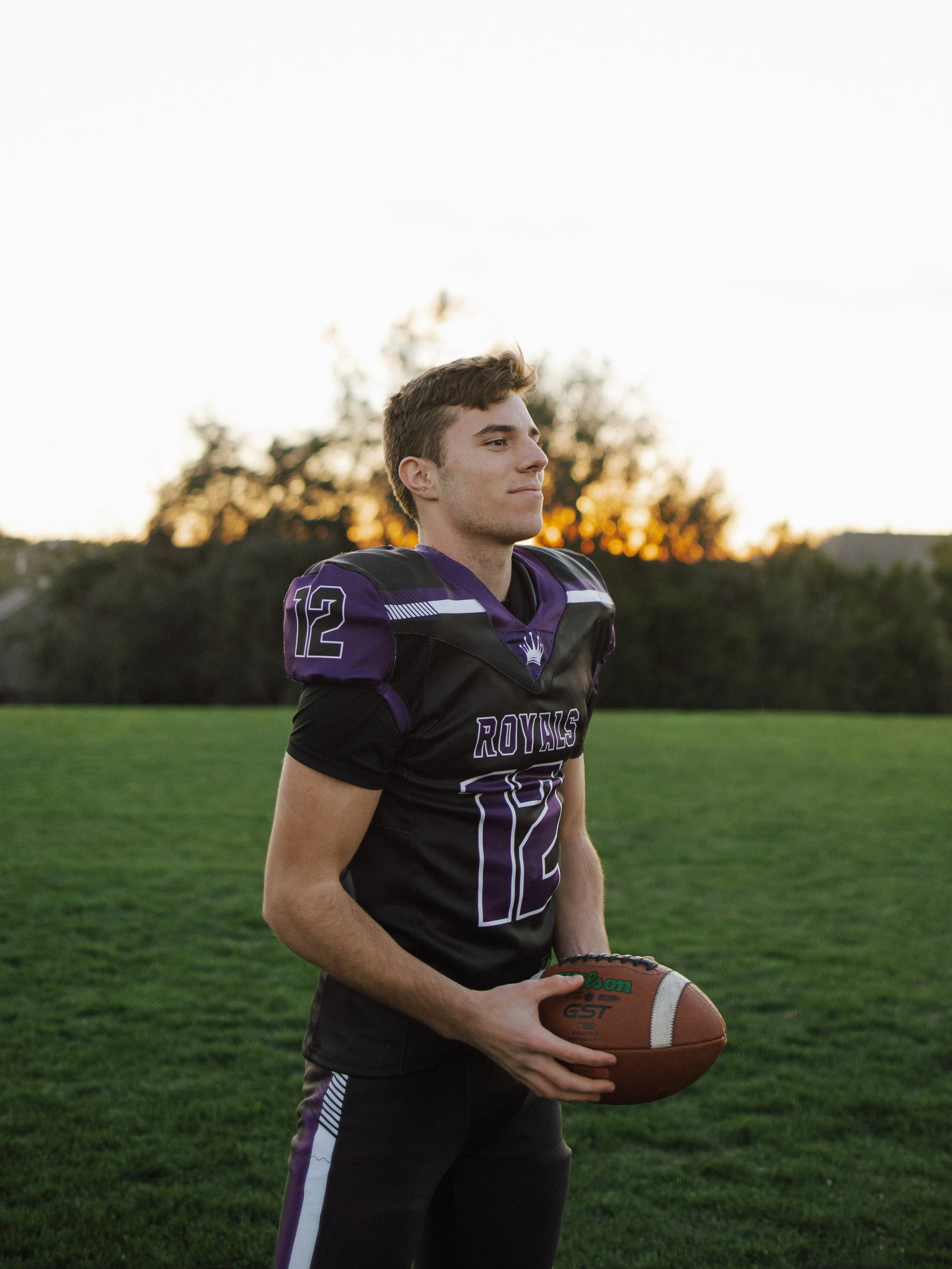 A young male football player standing on a field during sunset, holding a football, wearing a black and purple jersey with the number 12.
