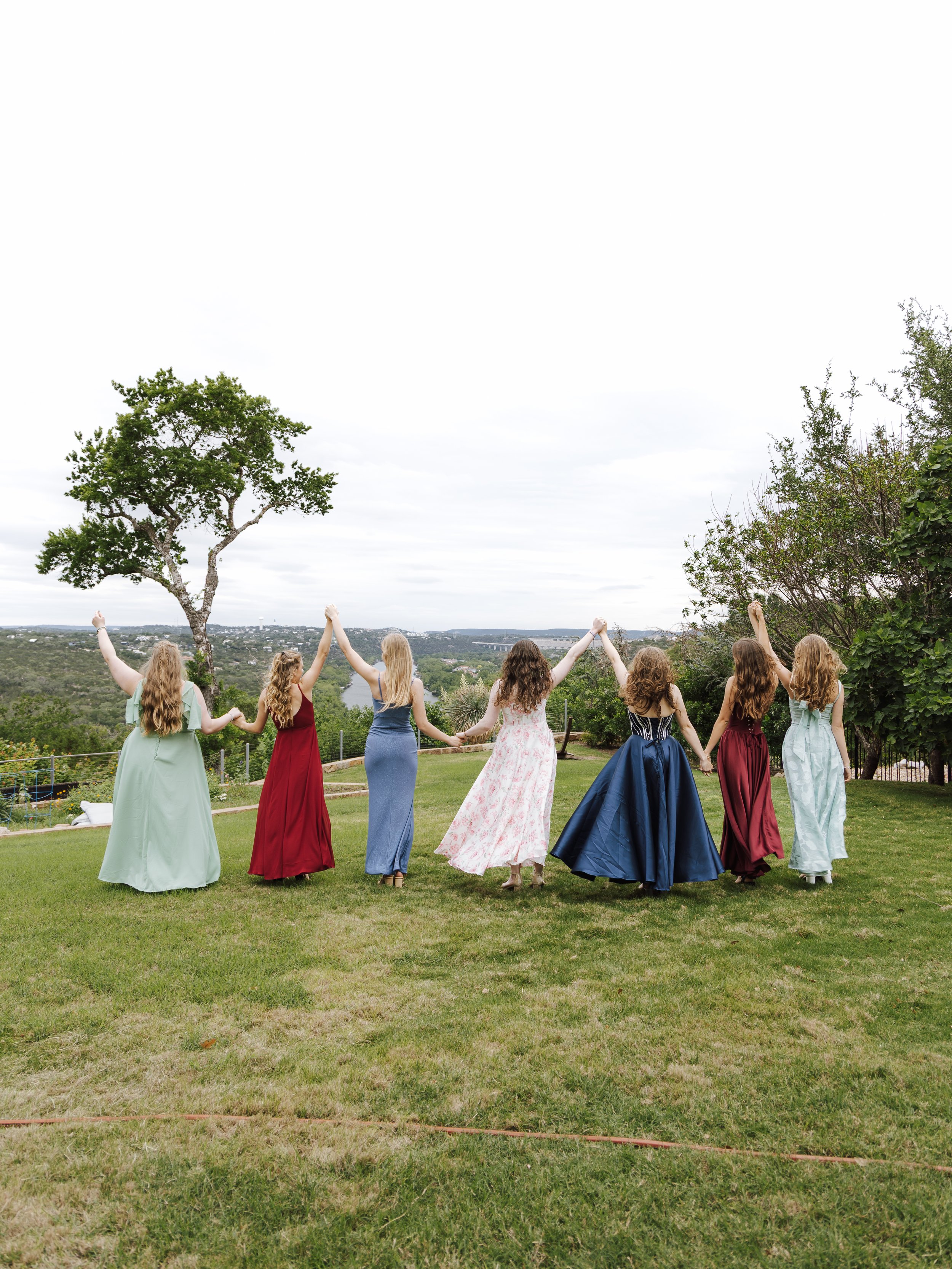 Seven women in colorful dresses holding hands and raising their arms in a circle on a grassy lawn with trees and a scenic view in the background.