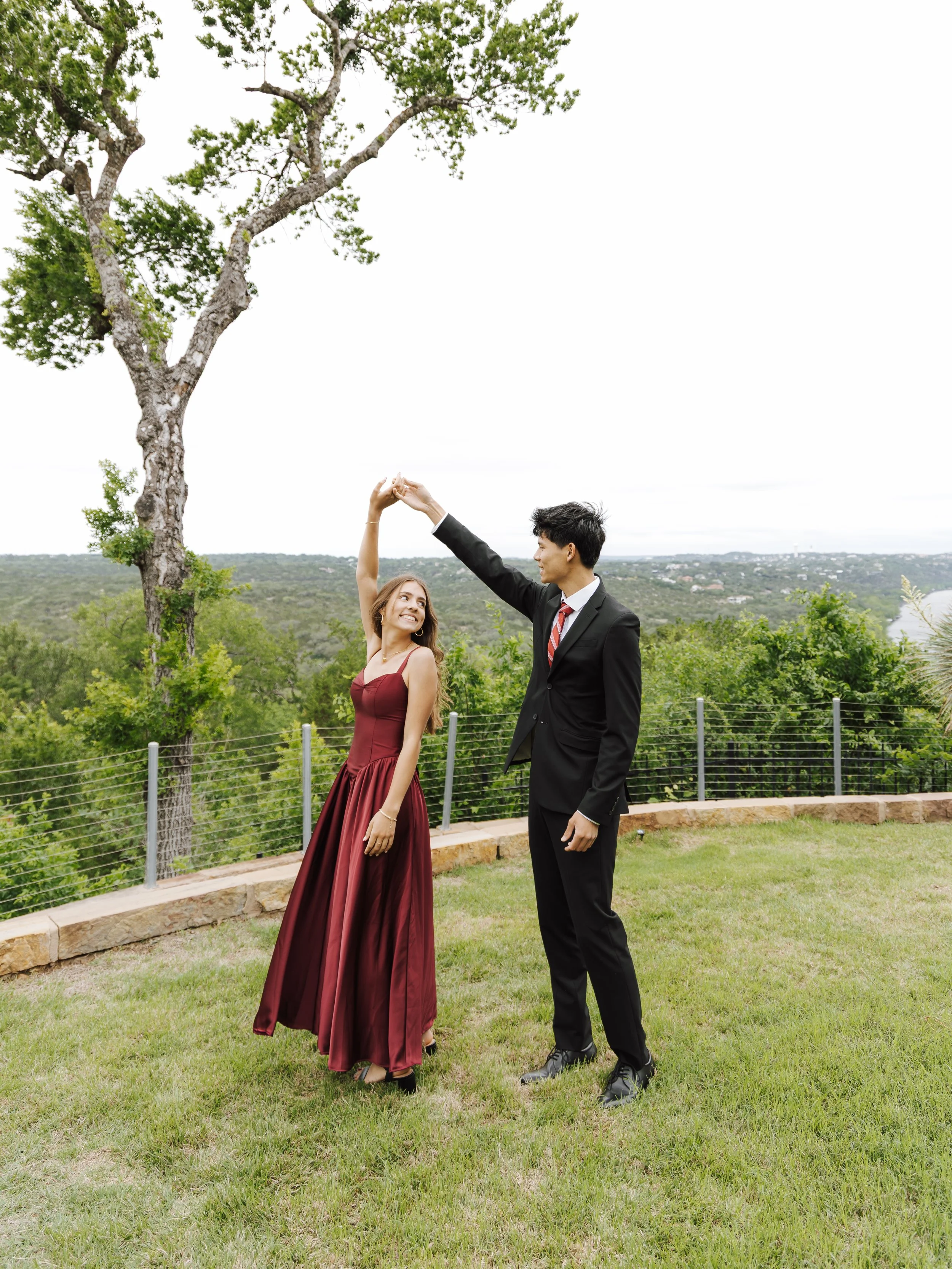 A young couple dancing outdoors on a grassy area with a railing, surrounded by trees and a scenic landscape, with the man wearing a black suit and the woman in a burgundy dress.