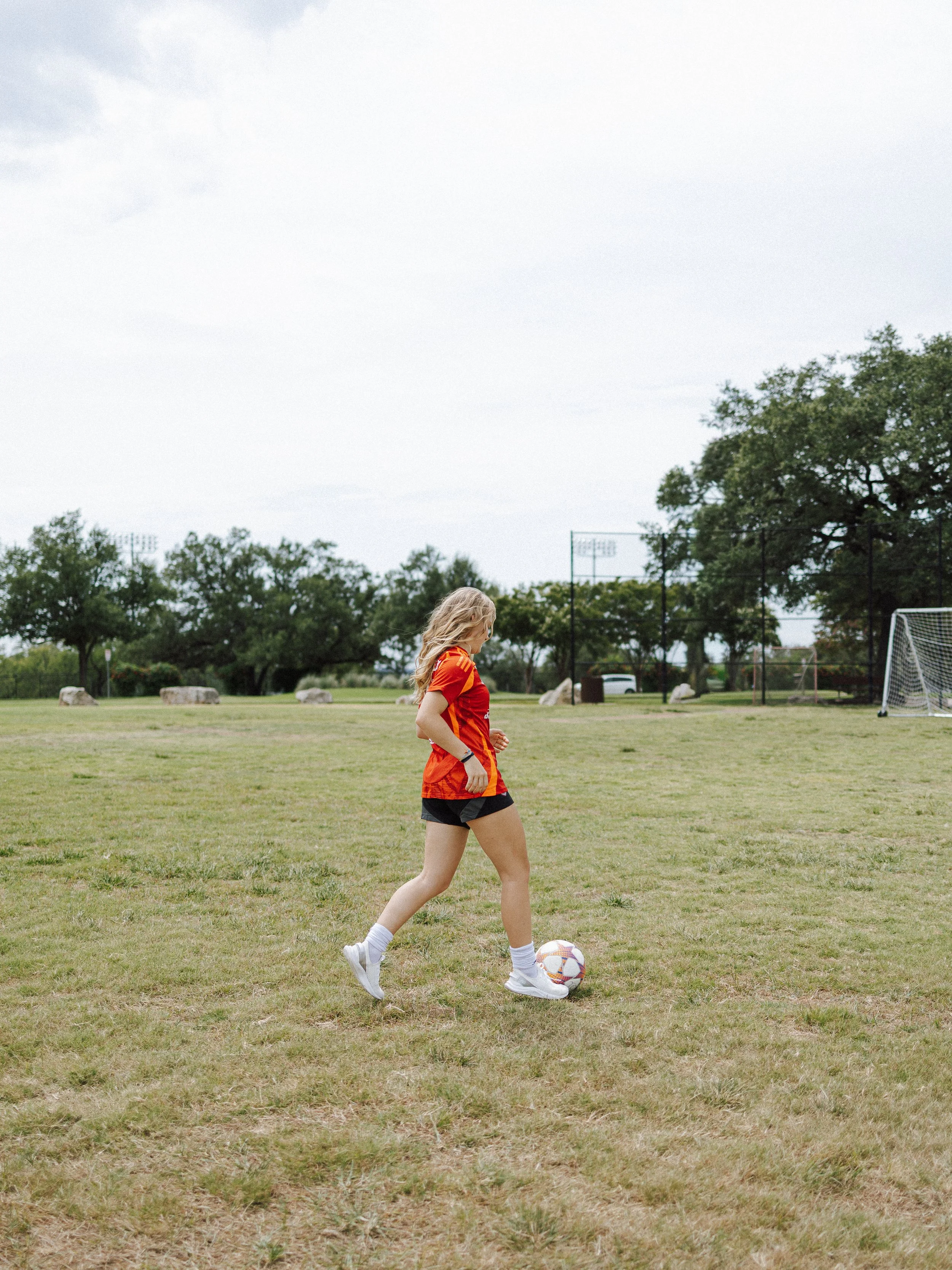 A young woman with blonde hair in a red sports jersey and black shorts is playing soccer on a grassy field. She is standing with a soccer ball at her feet, preparing to kick. There are trees and outdoor sports facilities in the background under an ov