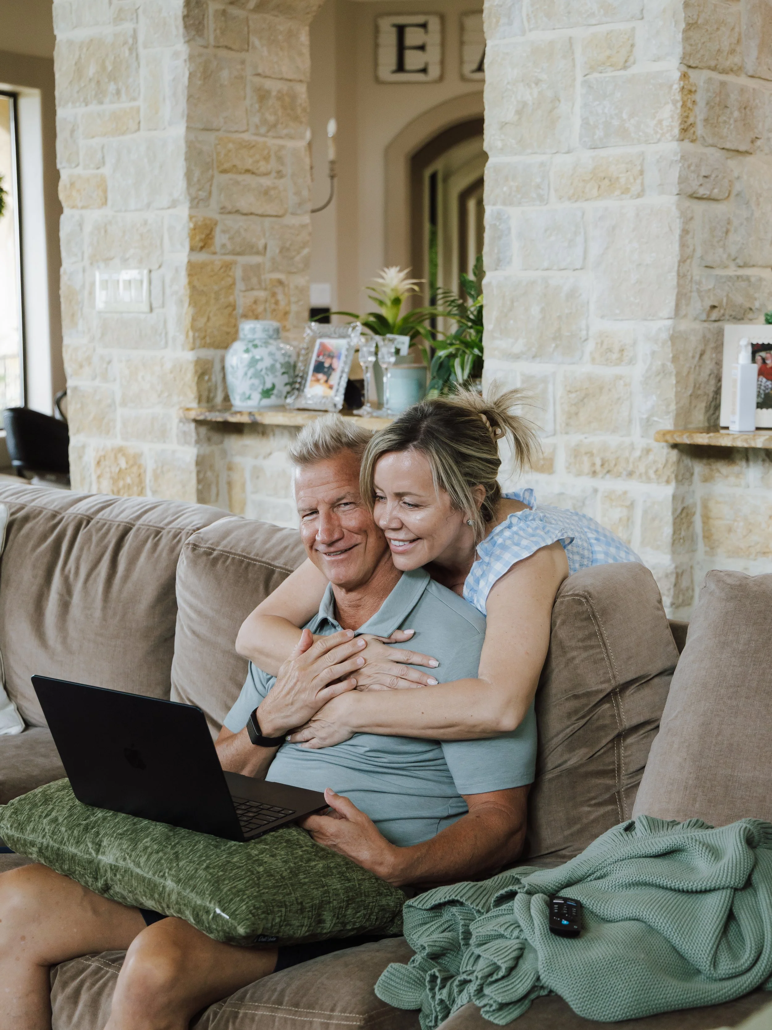 An elderly man and woman sitting on a beige sofa, watching a laptop screen, with the woman hugging the man from behind. The man has short gray hair, and the woman has blonde hair tied in a ponytail. There are various decorative items and framed photo