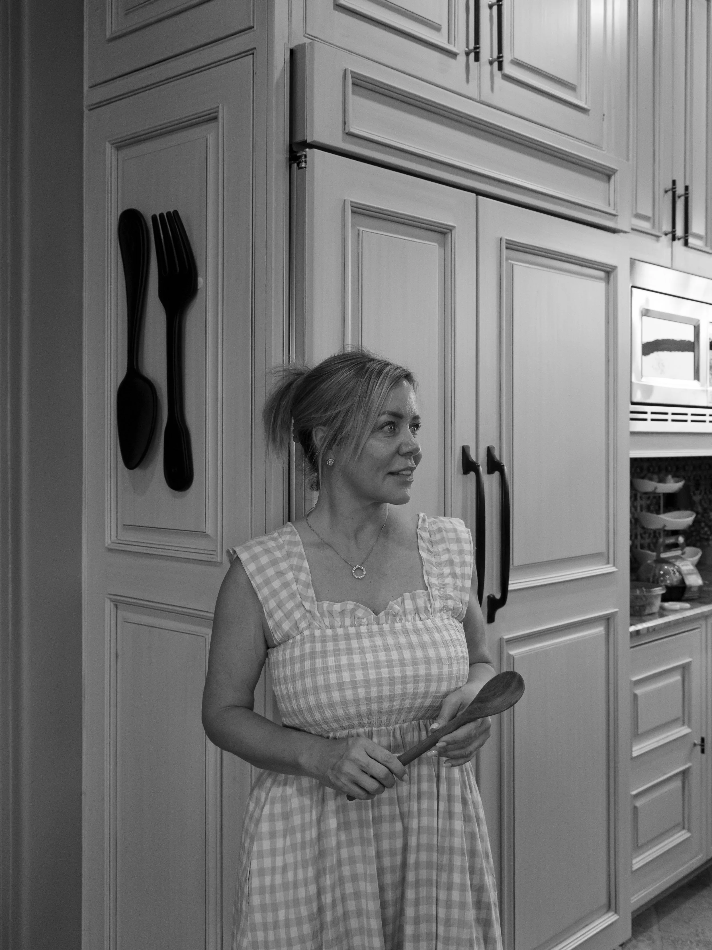 A woman in a checkered dress holding a wooden spoon standing in a kitchen with tall cabinets and utensils hanging on the wall.