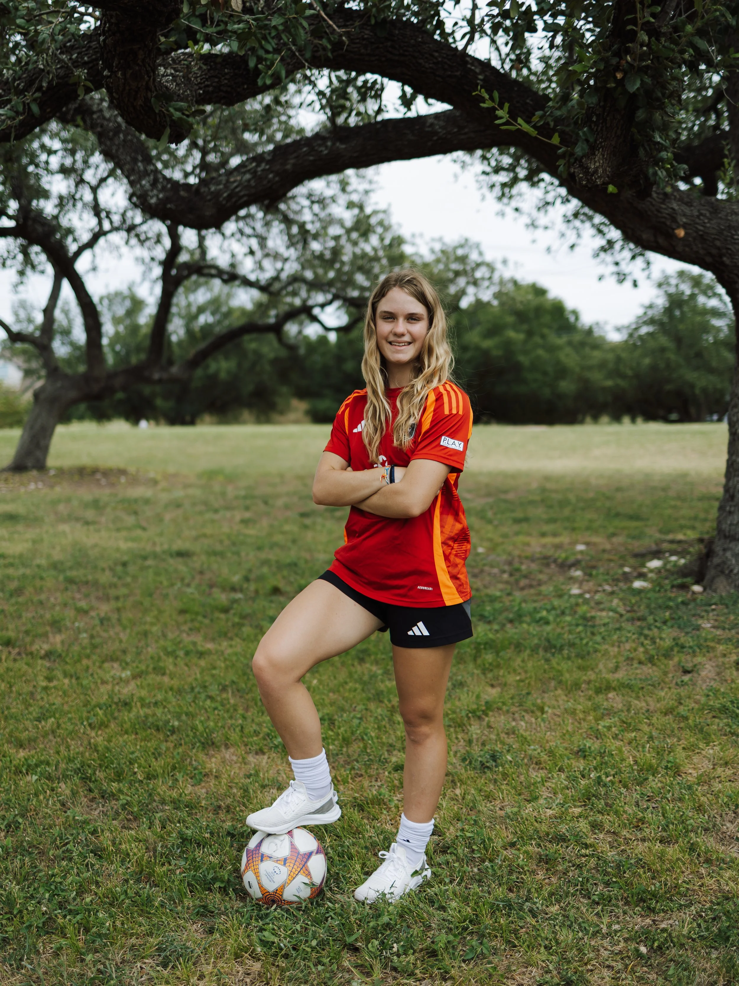 Young woman in sports uniform standing outdoors in a grassy field, with her arms crossed, one foot on a soccer ball, under trees with a clear sky behind her.