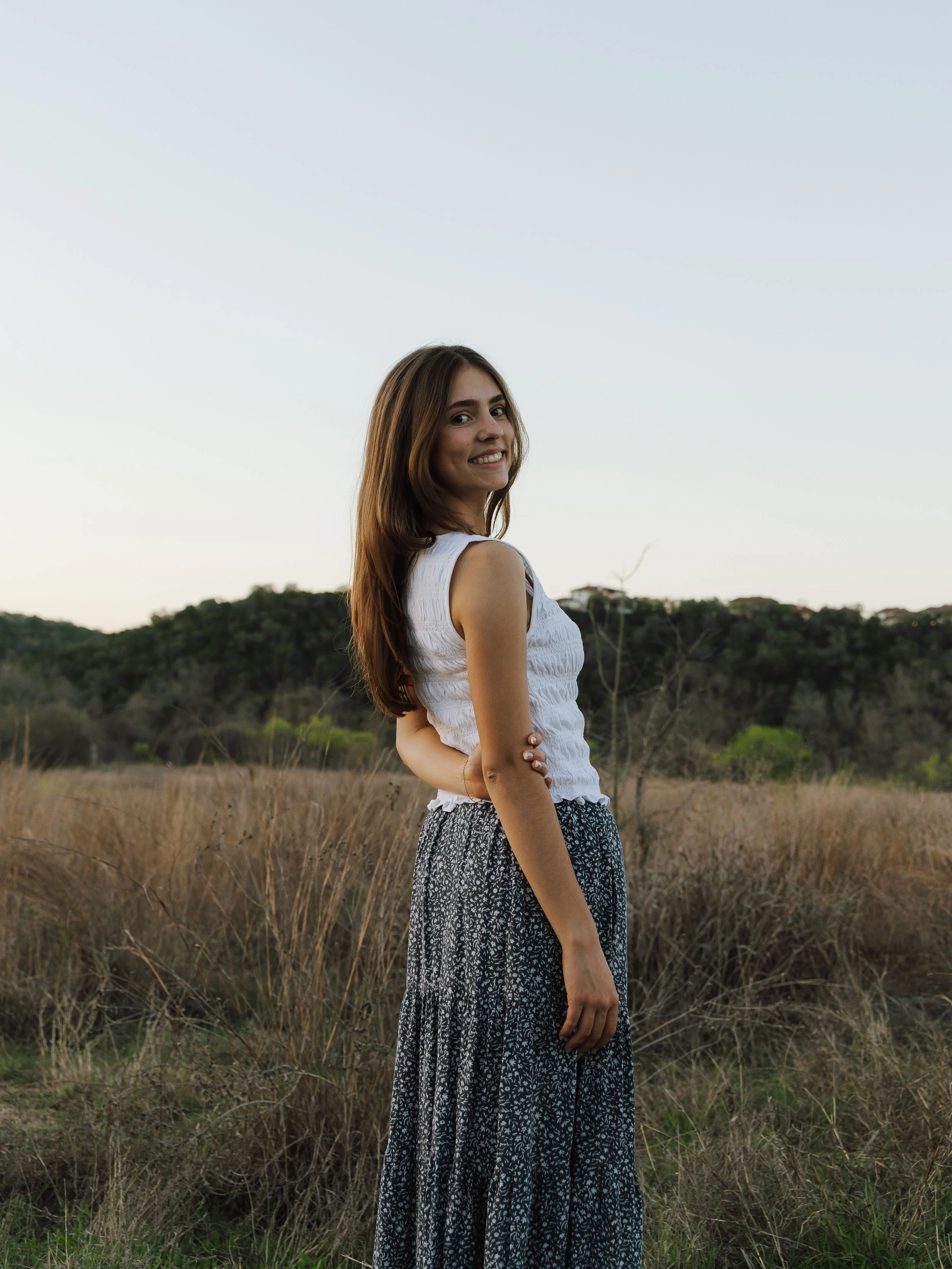 A young woman in a white sleeveless top and a long patterned skirt standing in a grassy field at dusk.