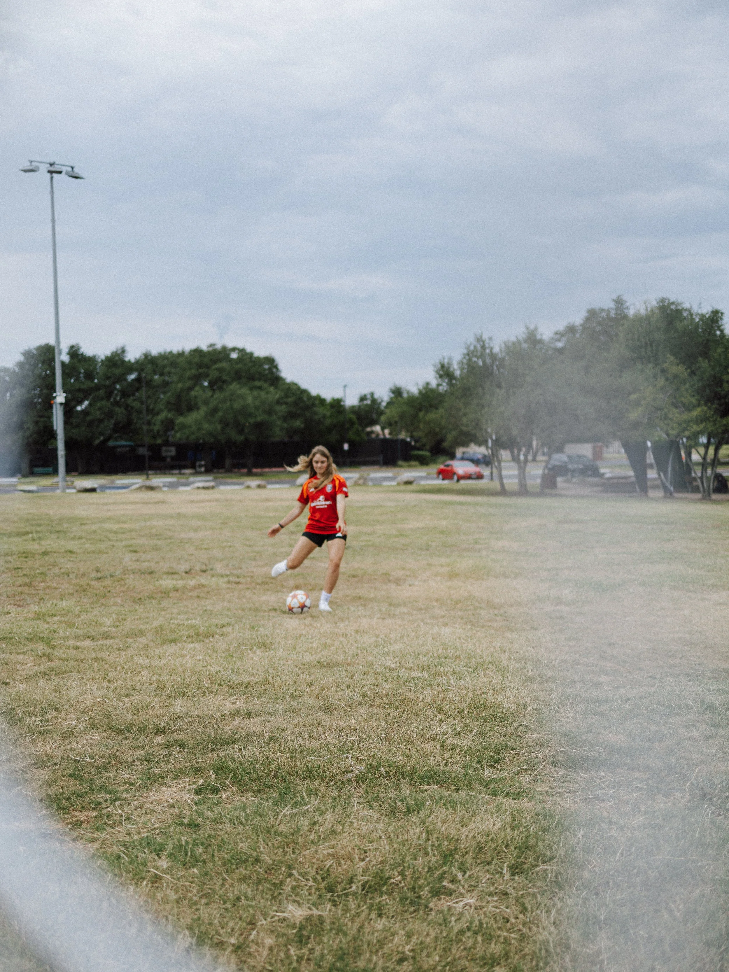 A young woman in a red sports jersey playing soccer on a grassy field during daytime, with a cloudy sky in the background and trees and parked cars in the distance.