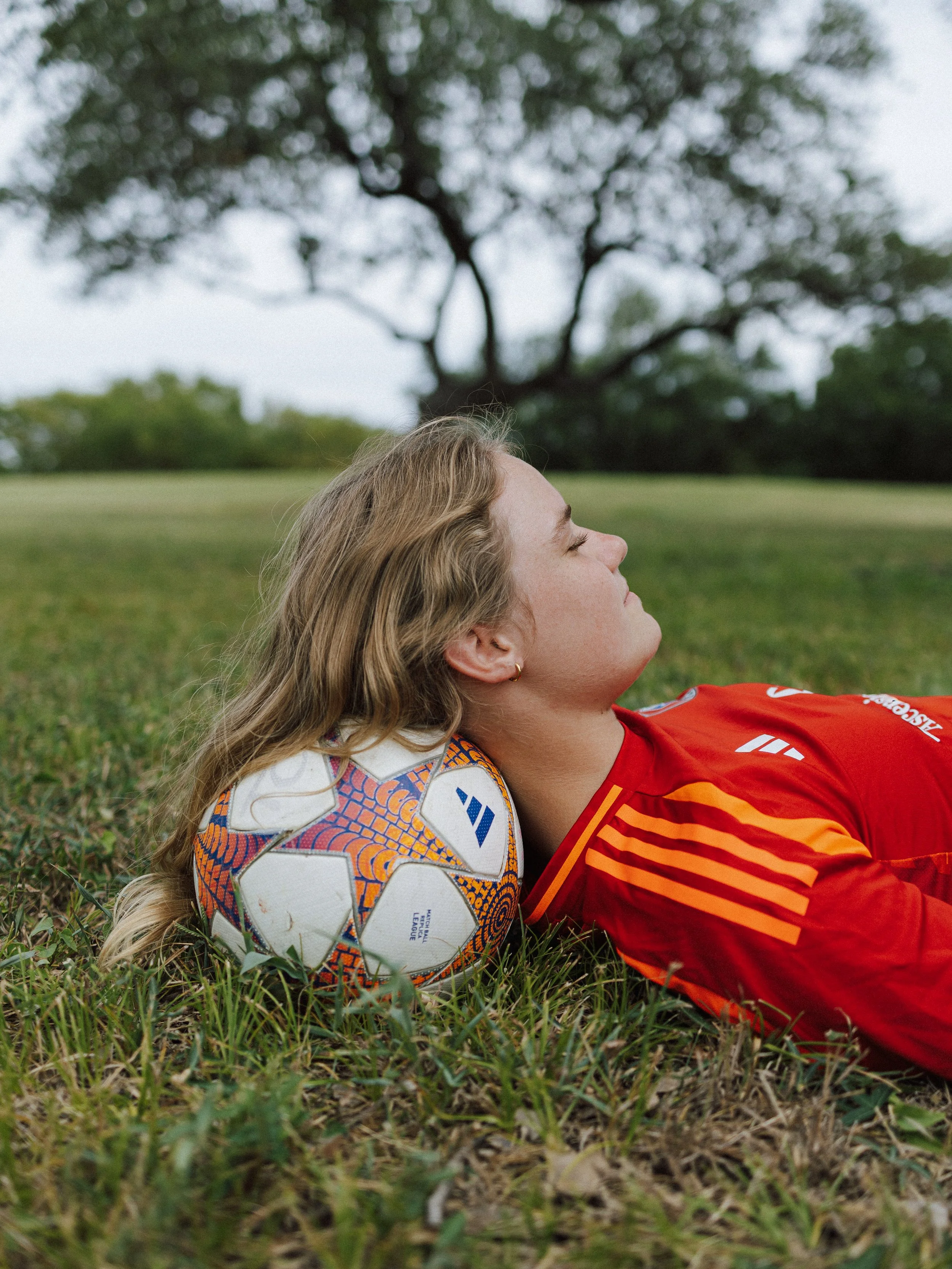 A woman with long blonde hair wearing a sweatshirt, lying on grass with eyes closed, resting her head against a soccer ball, outdoors in a park with a large tree in the background.