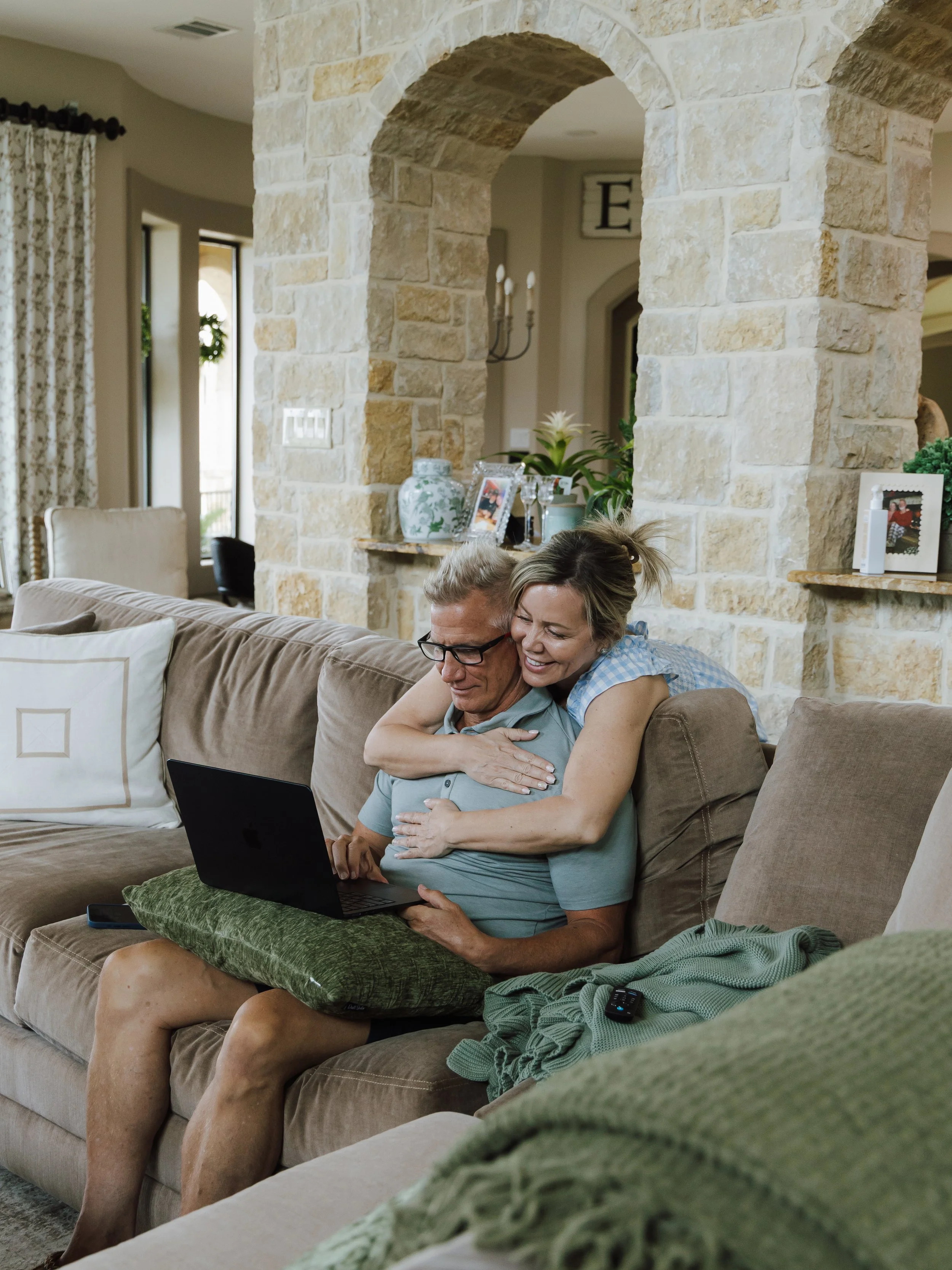A man and a woman sitting on a beige couch in a living room. The woman is hugging the man from behind while they look at a laptop. The room features a stone archway and mantel, with personal items and decorations on shelves and walls.