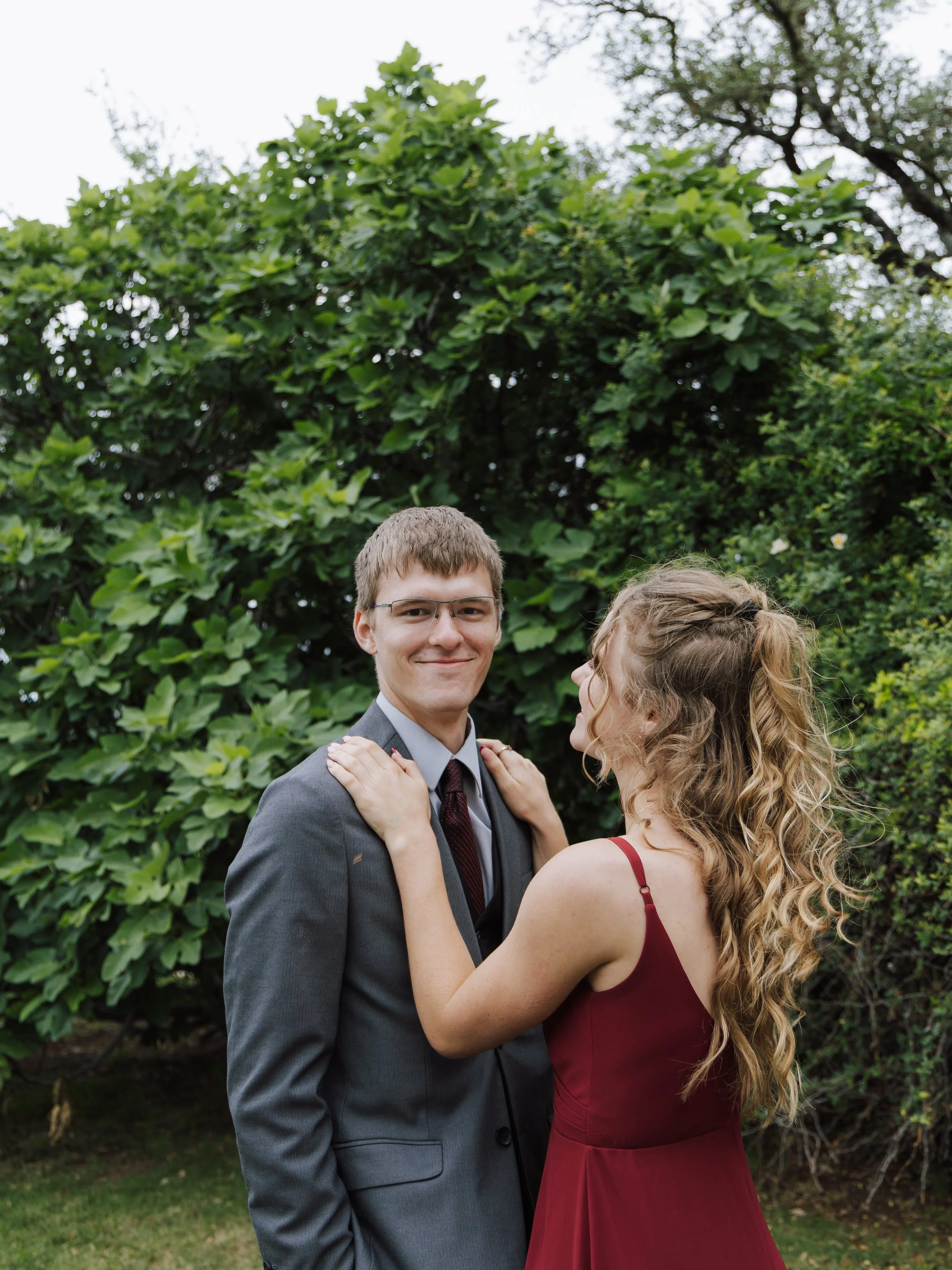 A young man in a gray suit smiling while a young woman with curly hair in a red dress touches his shoulders from the side, outdoors with green trees and bushes in the background.