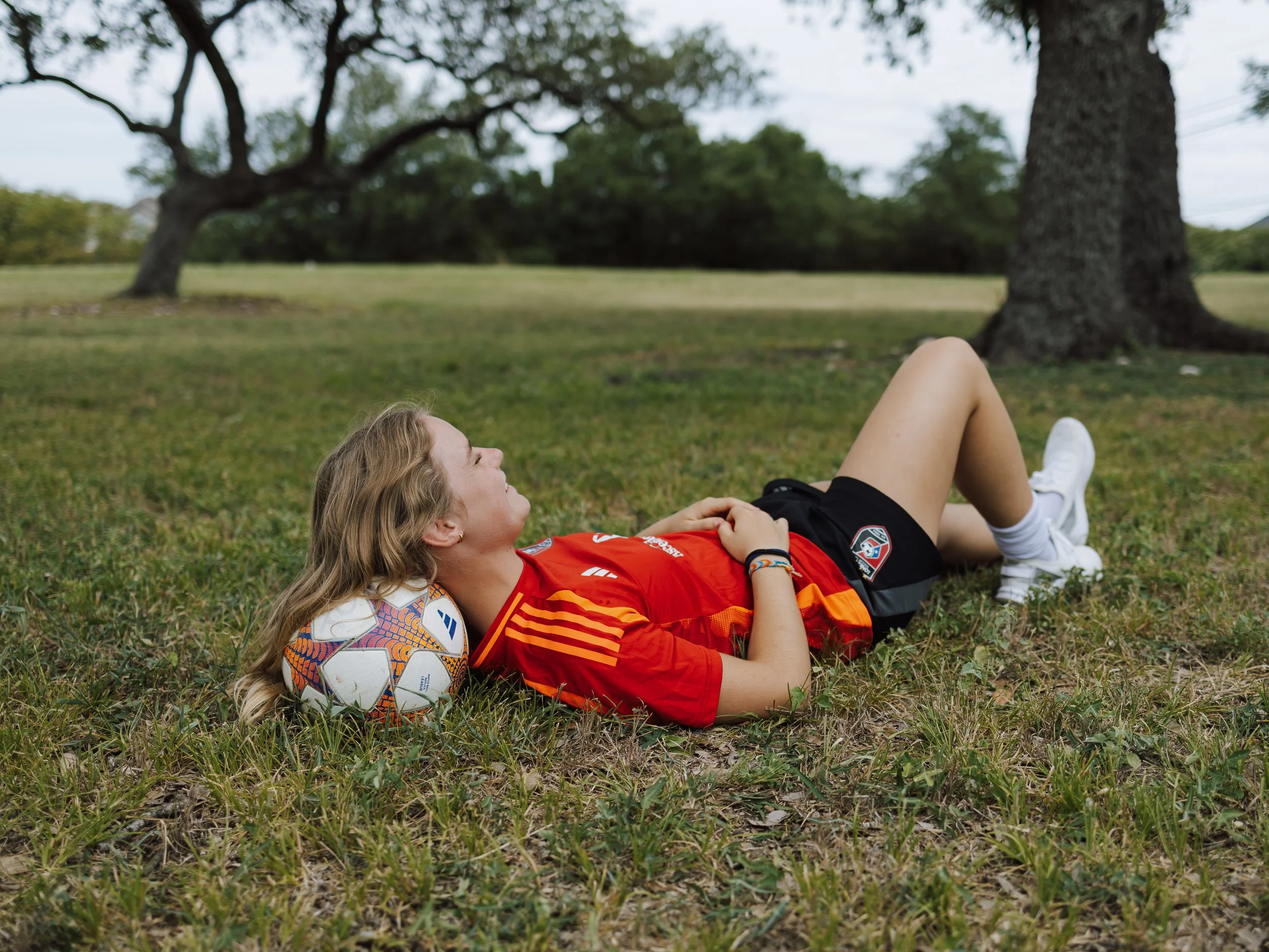 A young woman with long blonde hair lies on the grass, relaxing with a soccer ball near her head in a park with large trees in the background.