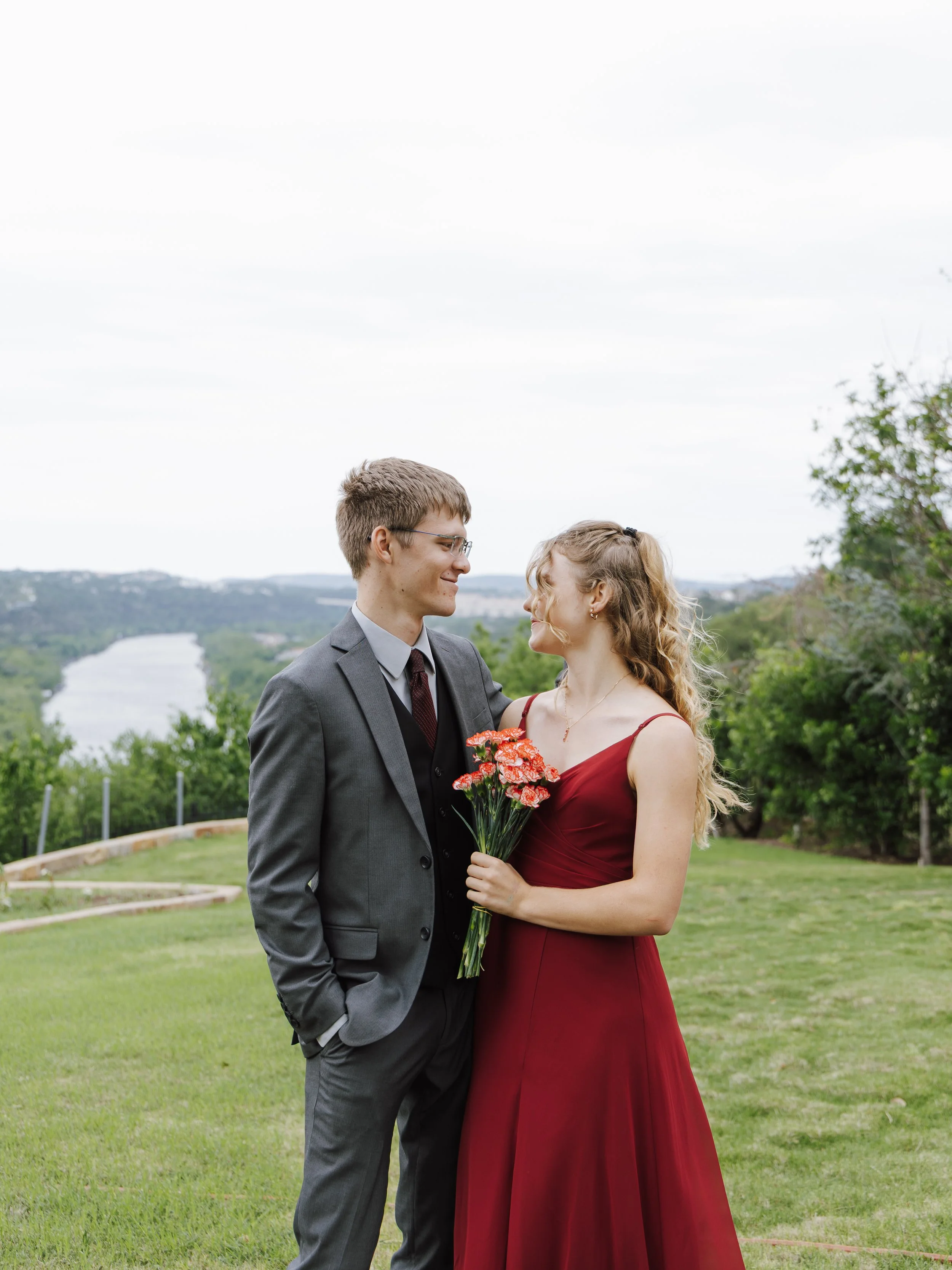 A couple standing outdoors on a grassy hill overlooking a river, with the woman holding a bouquet of pink flowers, both smiling at each other.