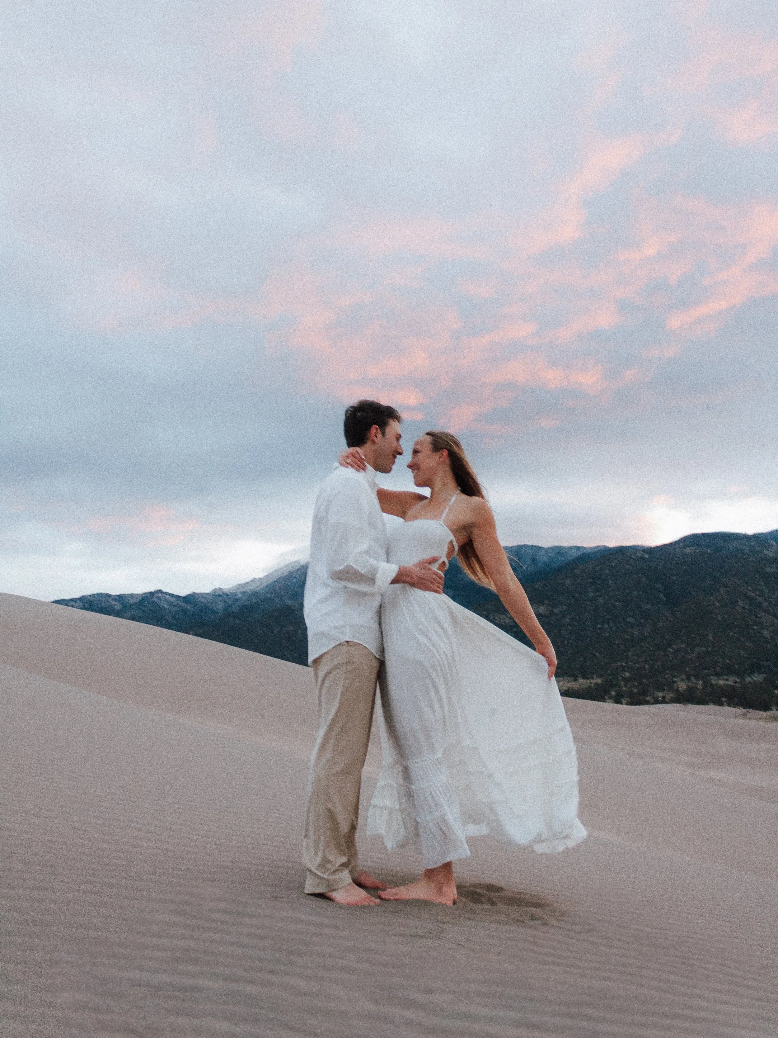 A couple in wedding attire embracing on a sandy desert with mountains in the background during sunset or sunrise.