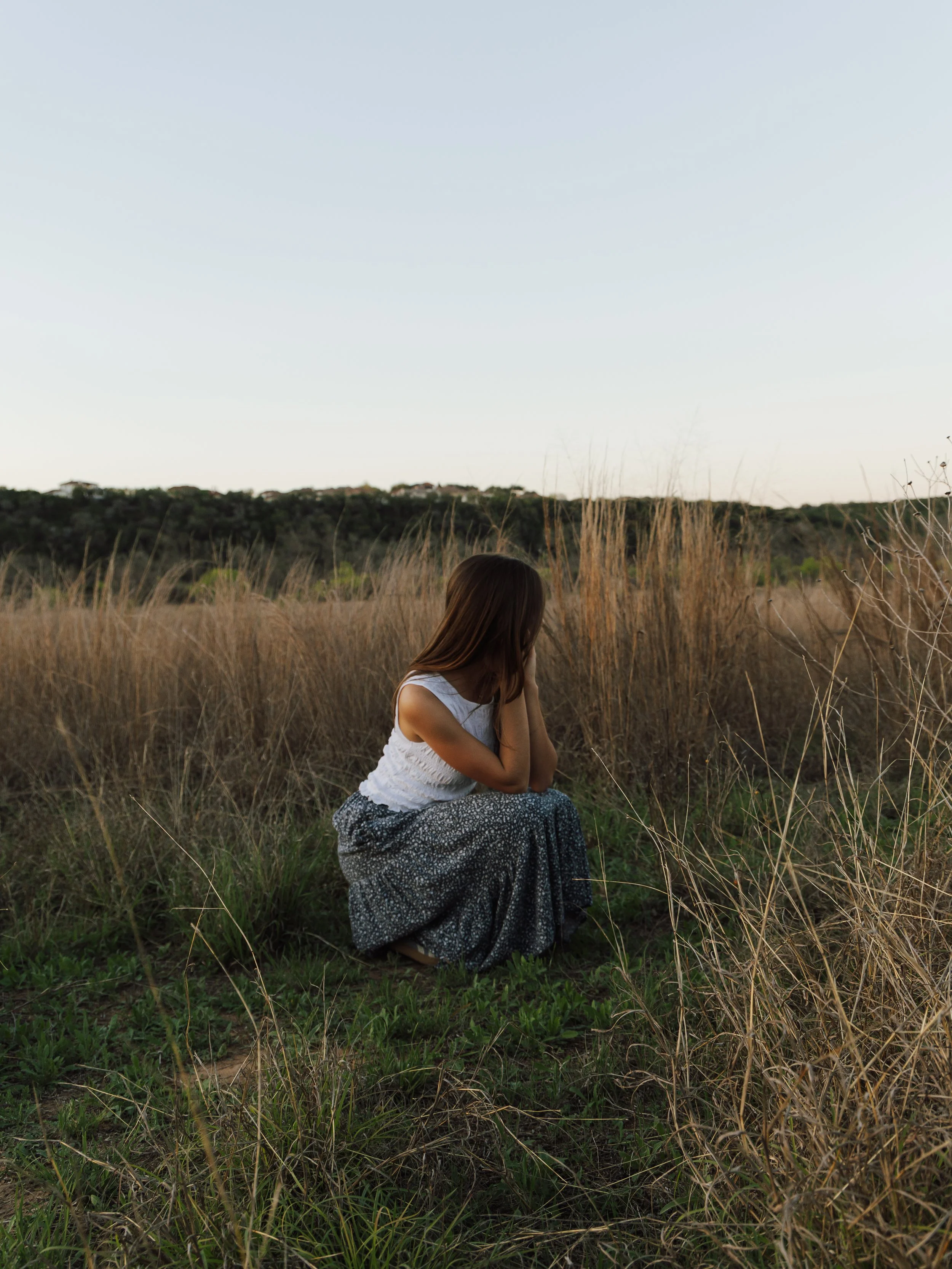 A woman kneeling in a grassy field with tall dry grass, her head bowed and her hands near her face, under a clear sky.