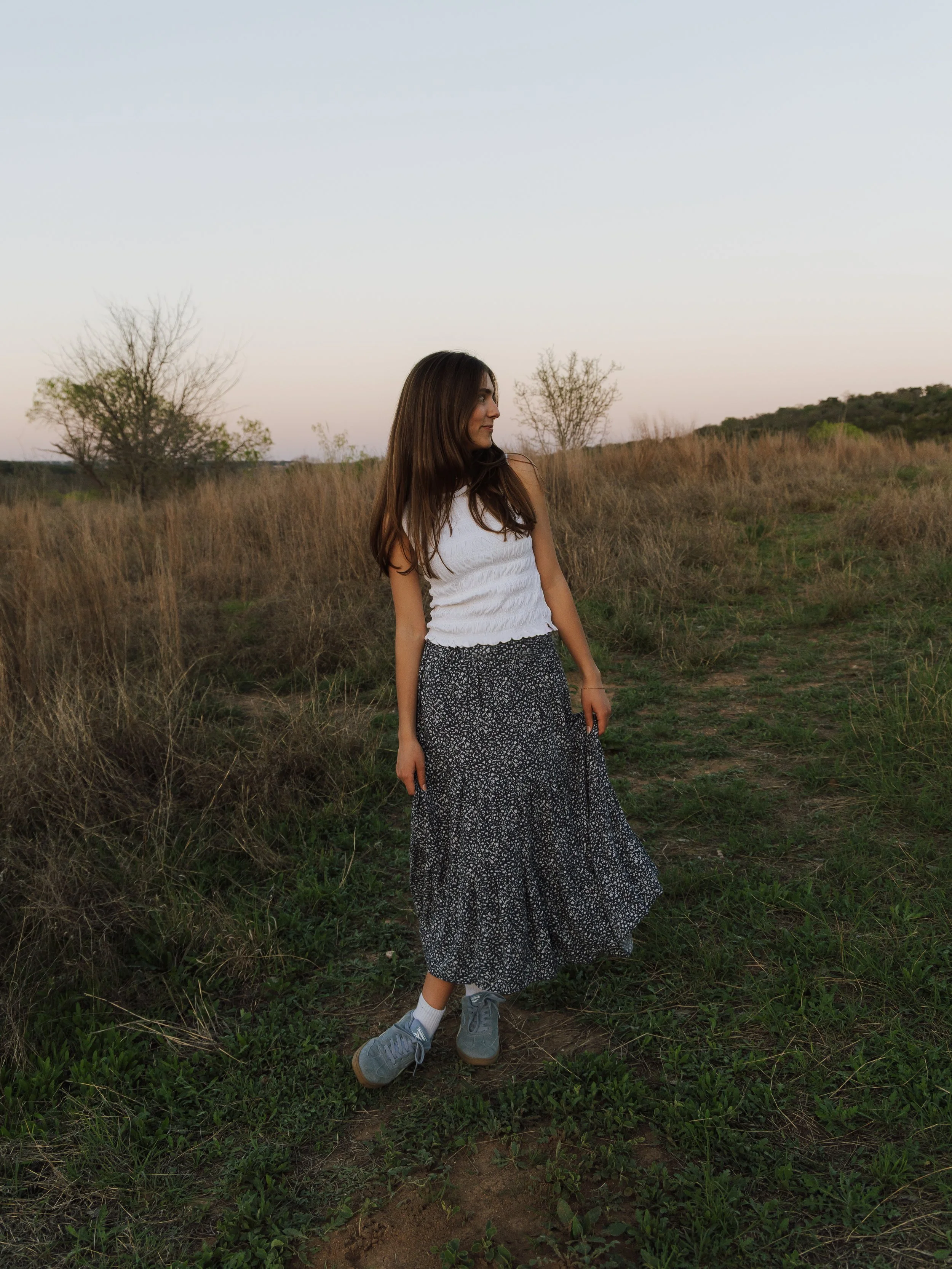 A young woman standing in a grassy field with dry bushes and trees, wearing a white sleeveless top, a long black and white patterned skirt, and gray sneakers, looking to her left against a clear sky at sunset.