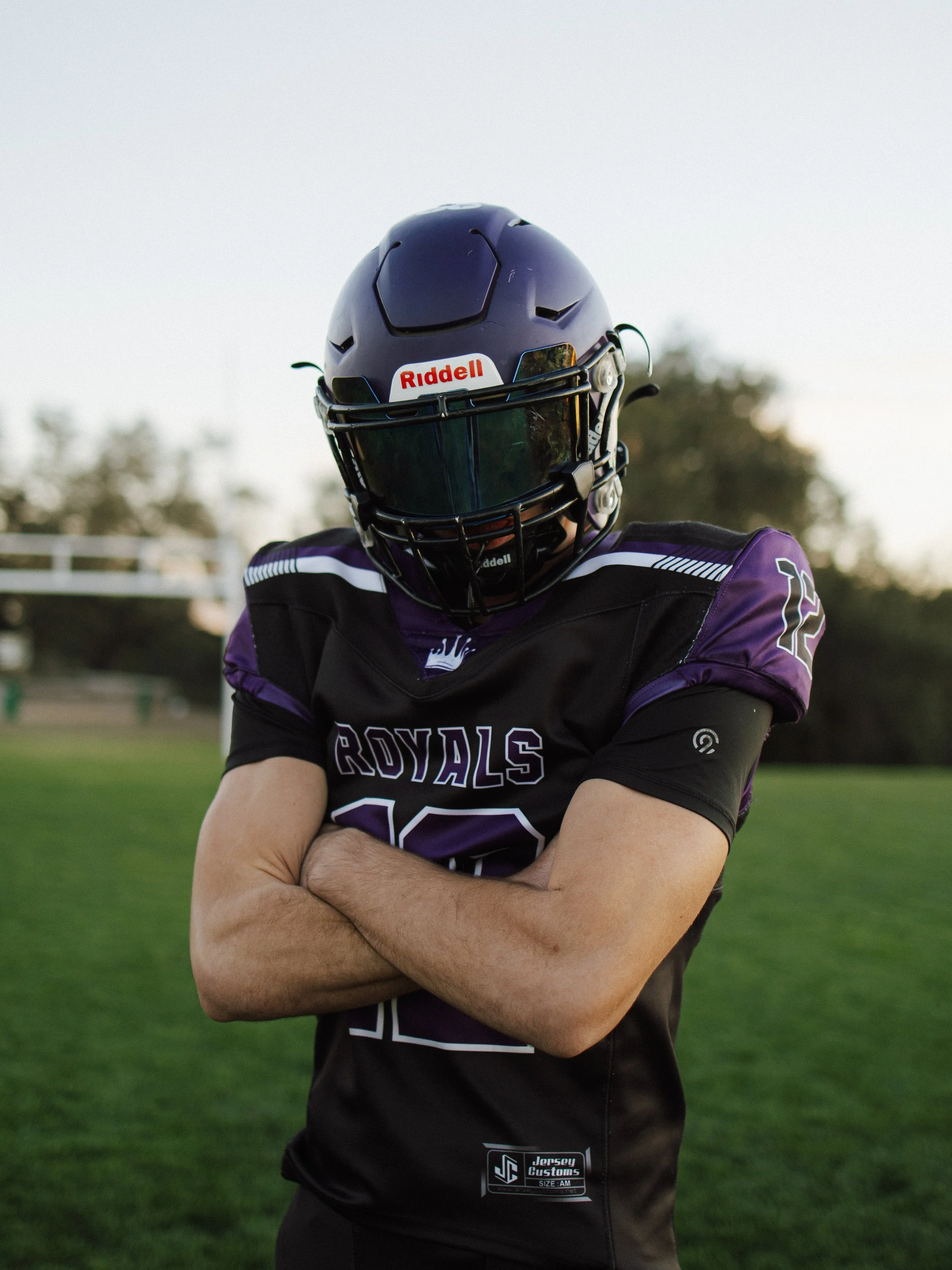 A football player in a purple and black jersey with crossed arms, wearing a dark helmet with a reflective visor on a grassy field.