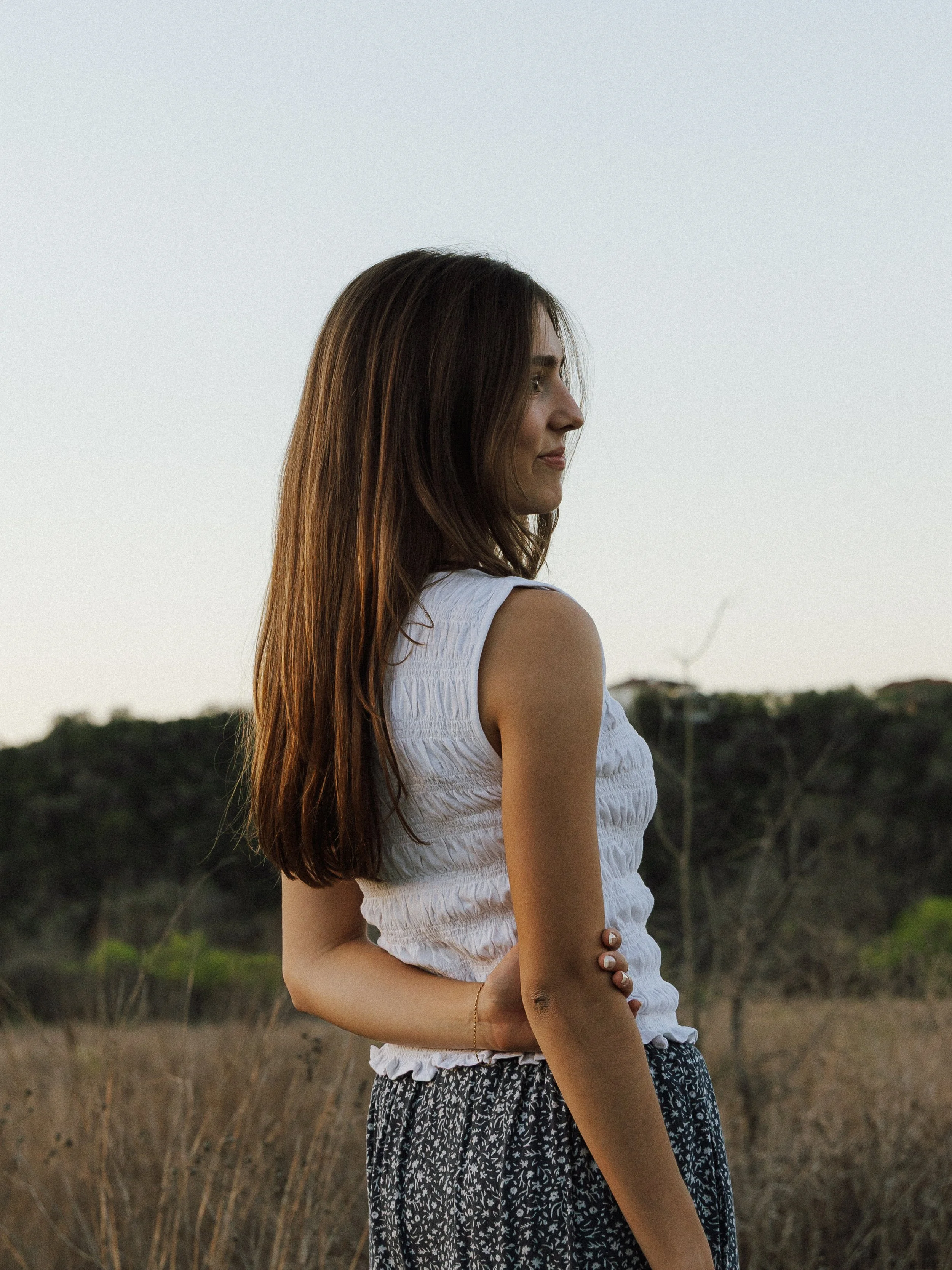Side profile of a woman with long brown hair standing outdoors in a field with dry grass and distant trees under a clear sky.