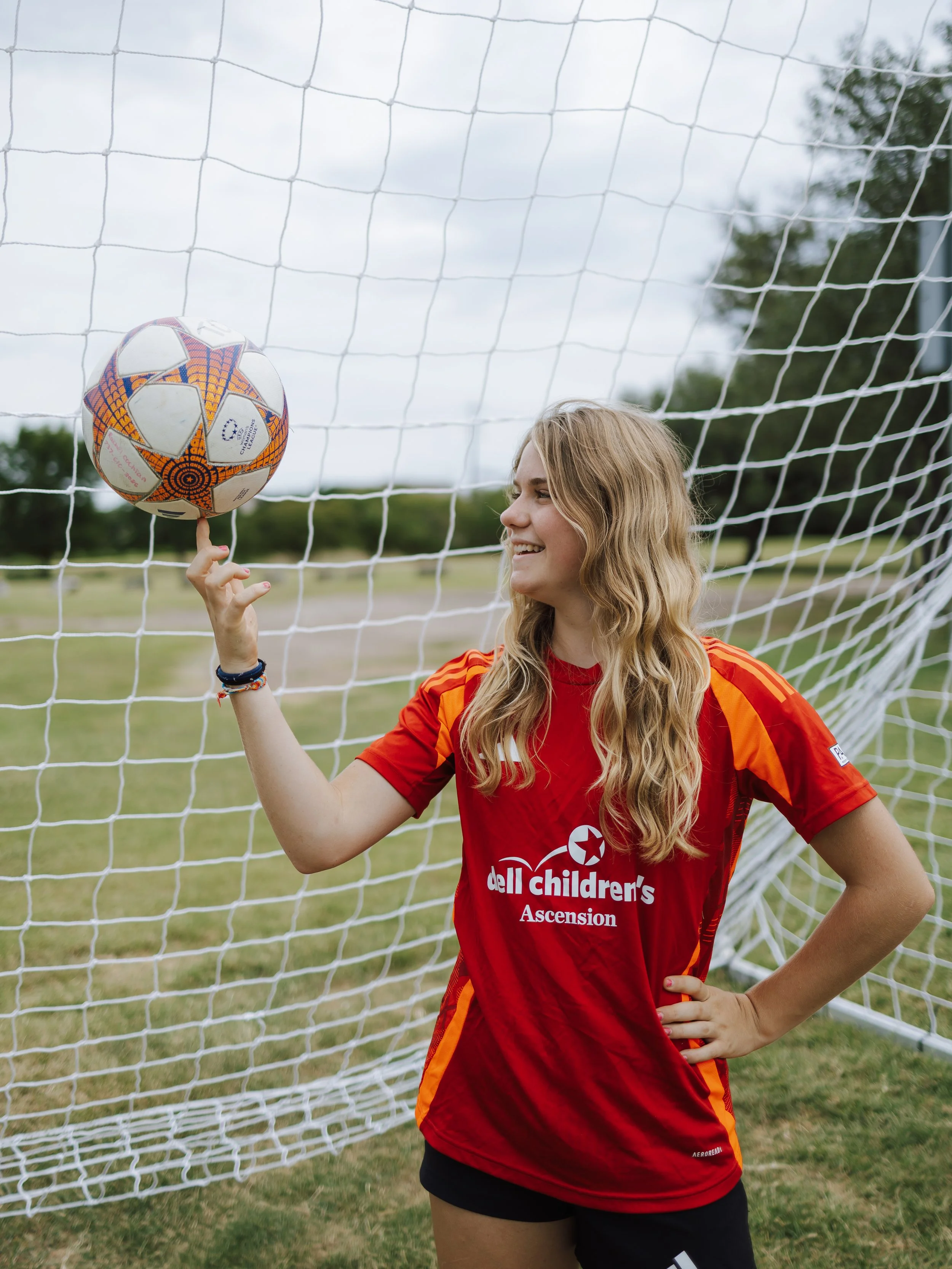 A young woman with long blonde hair is standing in front of a soccer goal net on a grassy field. She is wearing a red and orange sports jersey with the logo of 'dell children's Ascension' and black shorts. She is smiling and spinning a soccer ball on
