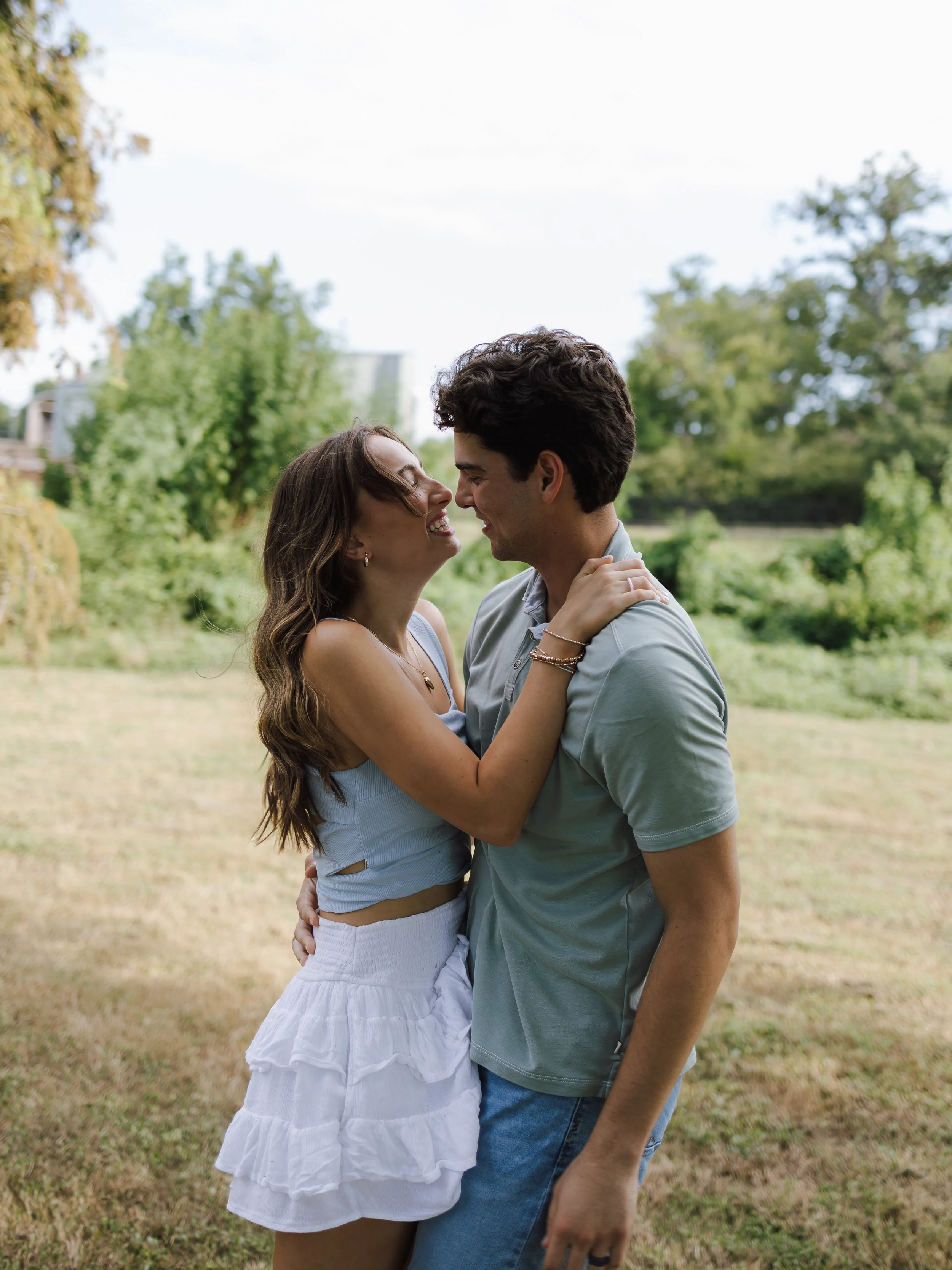 A couple smiling and embracing outdoors in a park with trees in the background.