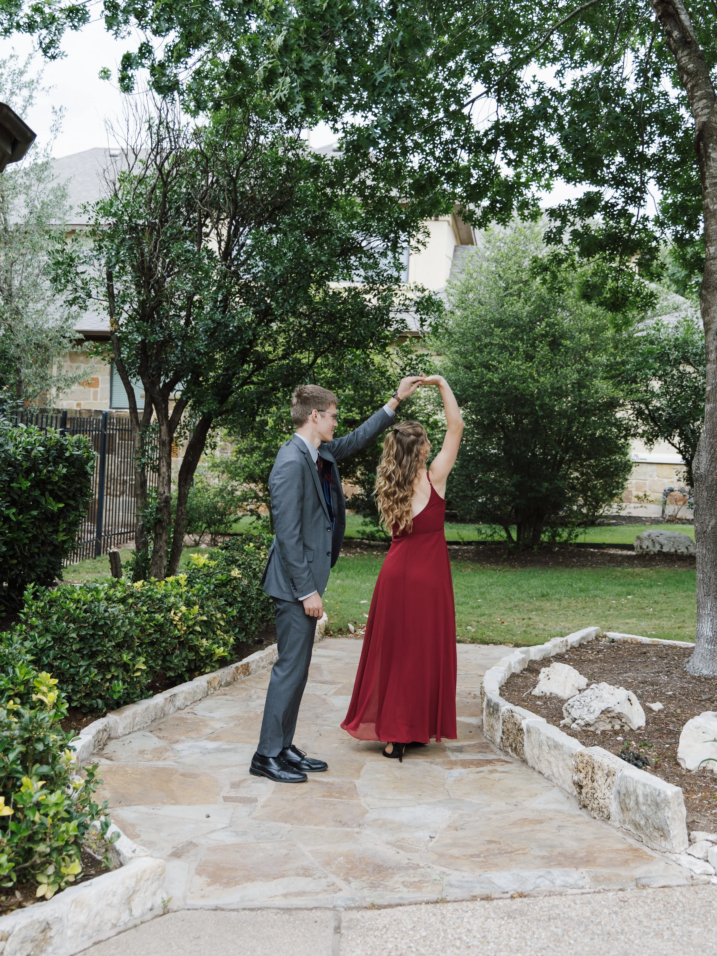 A young man in a gray suit is dancing with a young woman in a red dress on a stone pathway, surrounded by trees and greenery.
