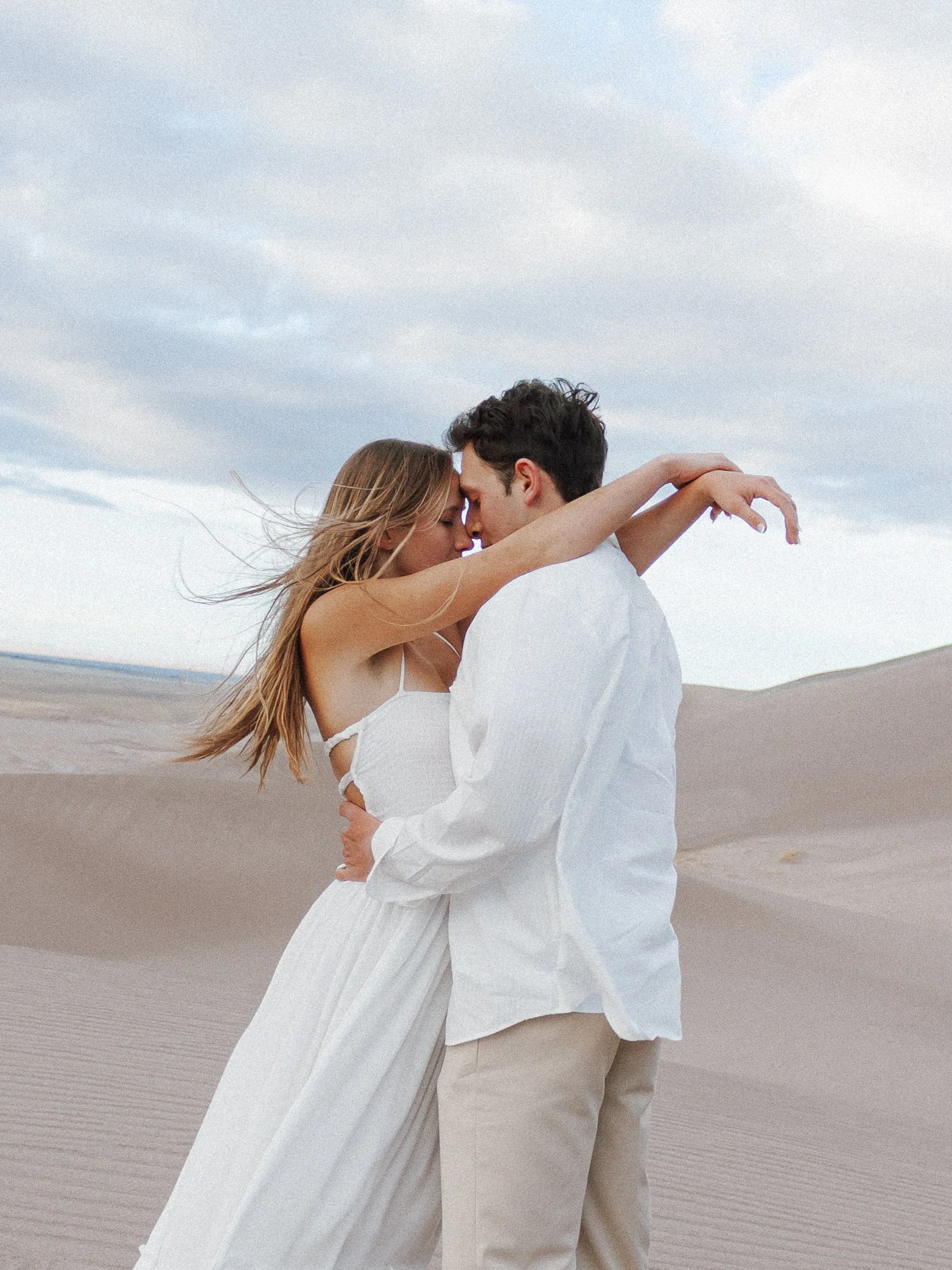 A couple embracing on a sandy beach with waves in the background, both dressed in white, with the woman wearing a flowing dress and the man in a white shirt. They are holding each other closely, with their foreheads touching.
