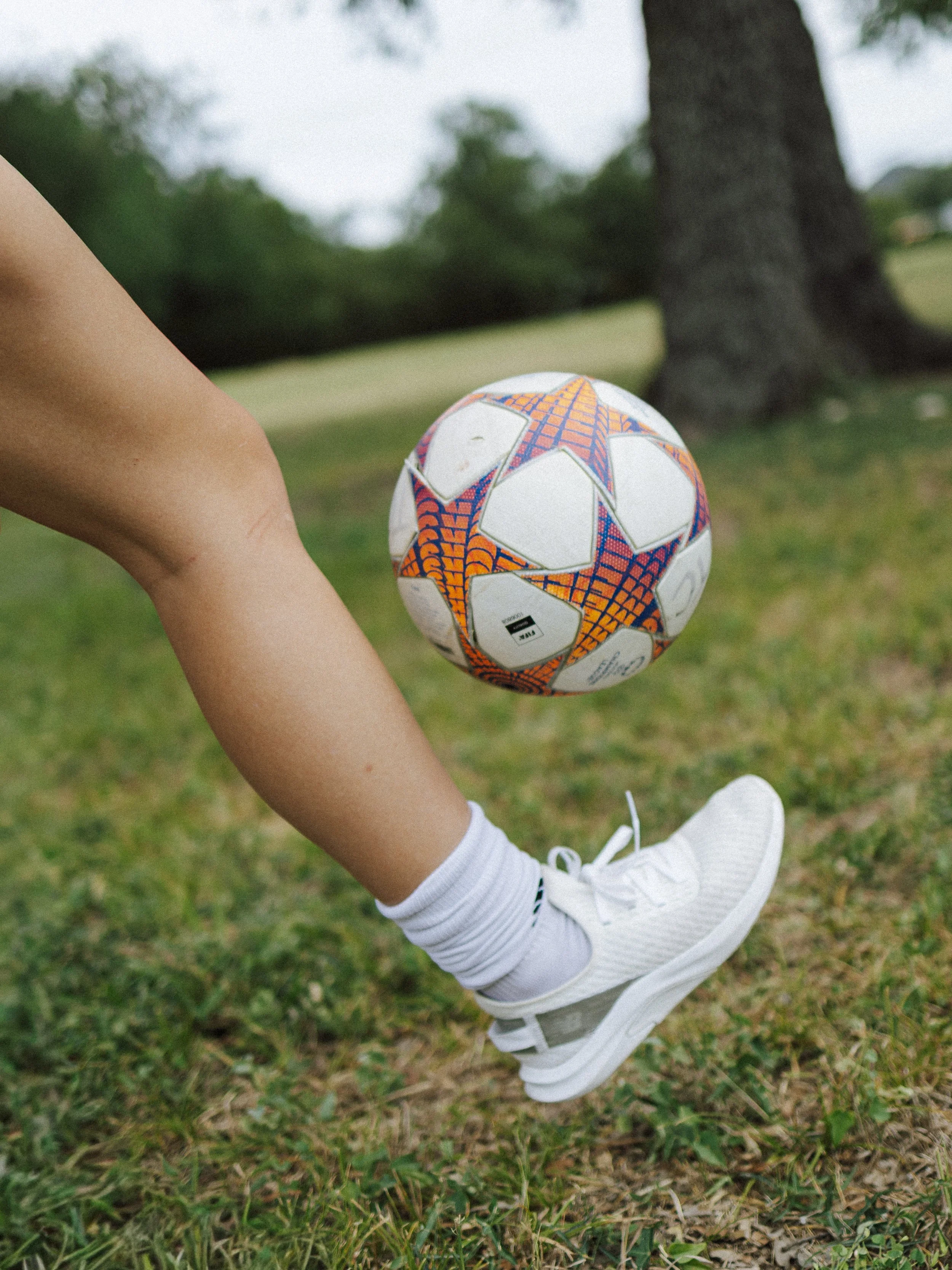 A person wearing white sneakers, white socks, and a white skirt is kicking a soccer ball with a colorful orange, blue, and white pattern on a grassy field with trees in the background.