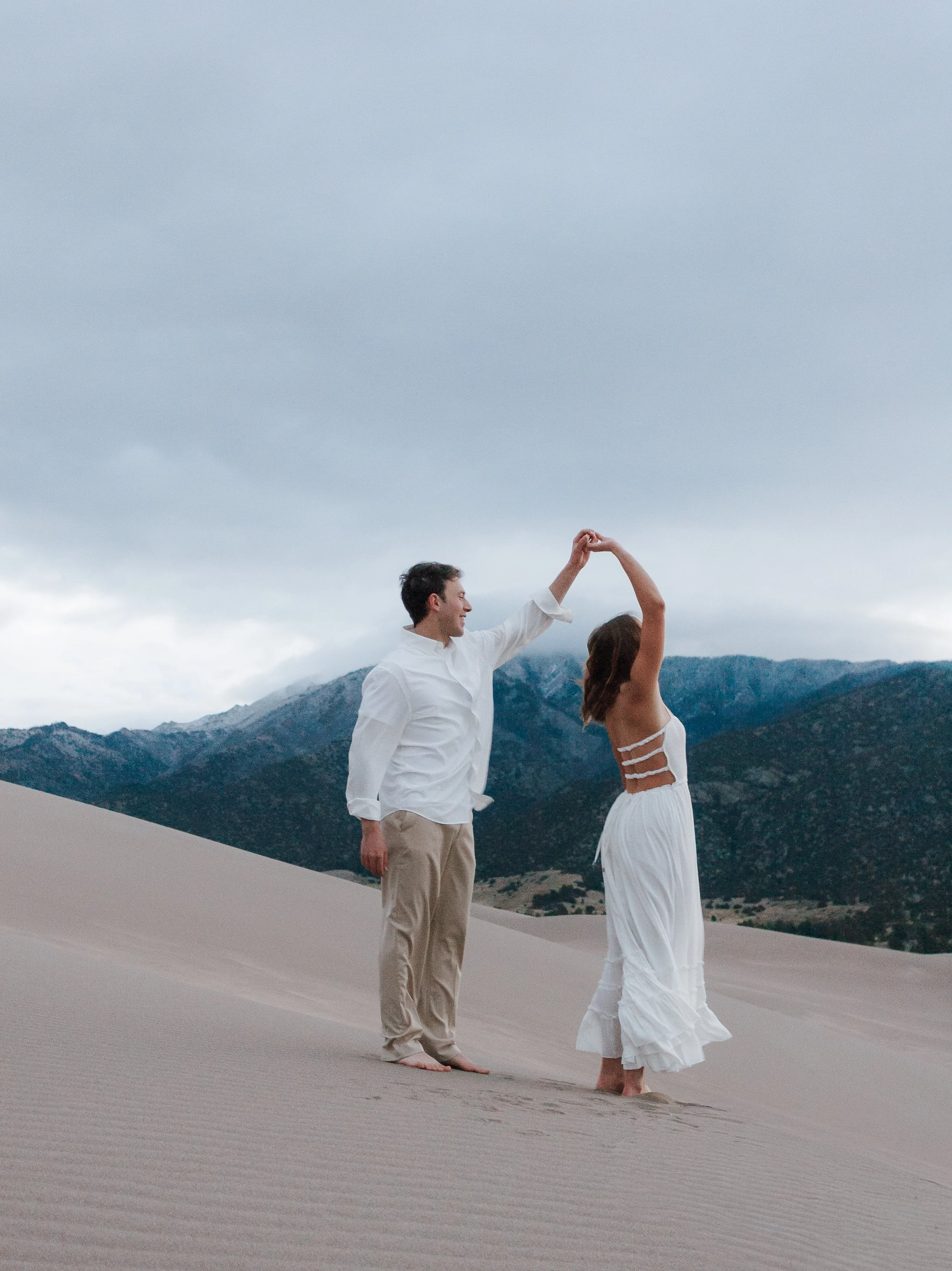 A couple dancing on sand dunes with mountains in the background under cloudy skies.