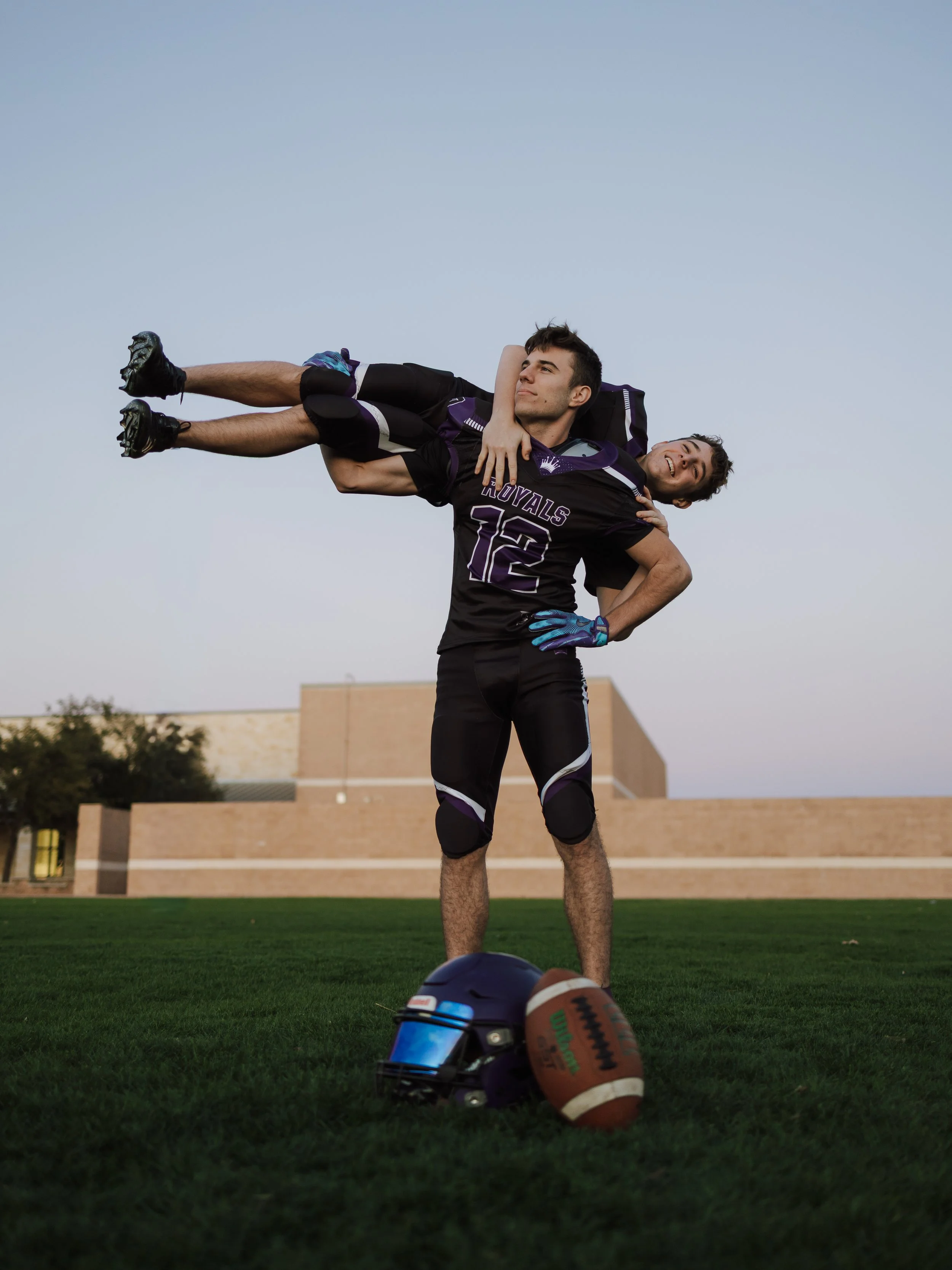 Two young men in sports gear, one standing with hand on hip, the other lying across shoulders of the standing man, on a grassy field with a football helmet and football in the foreground, during evening or early morning.