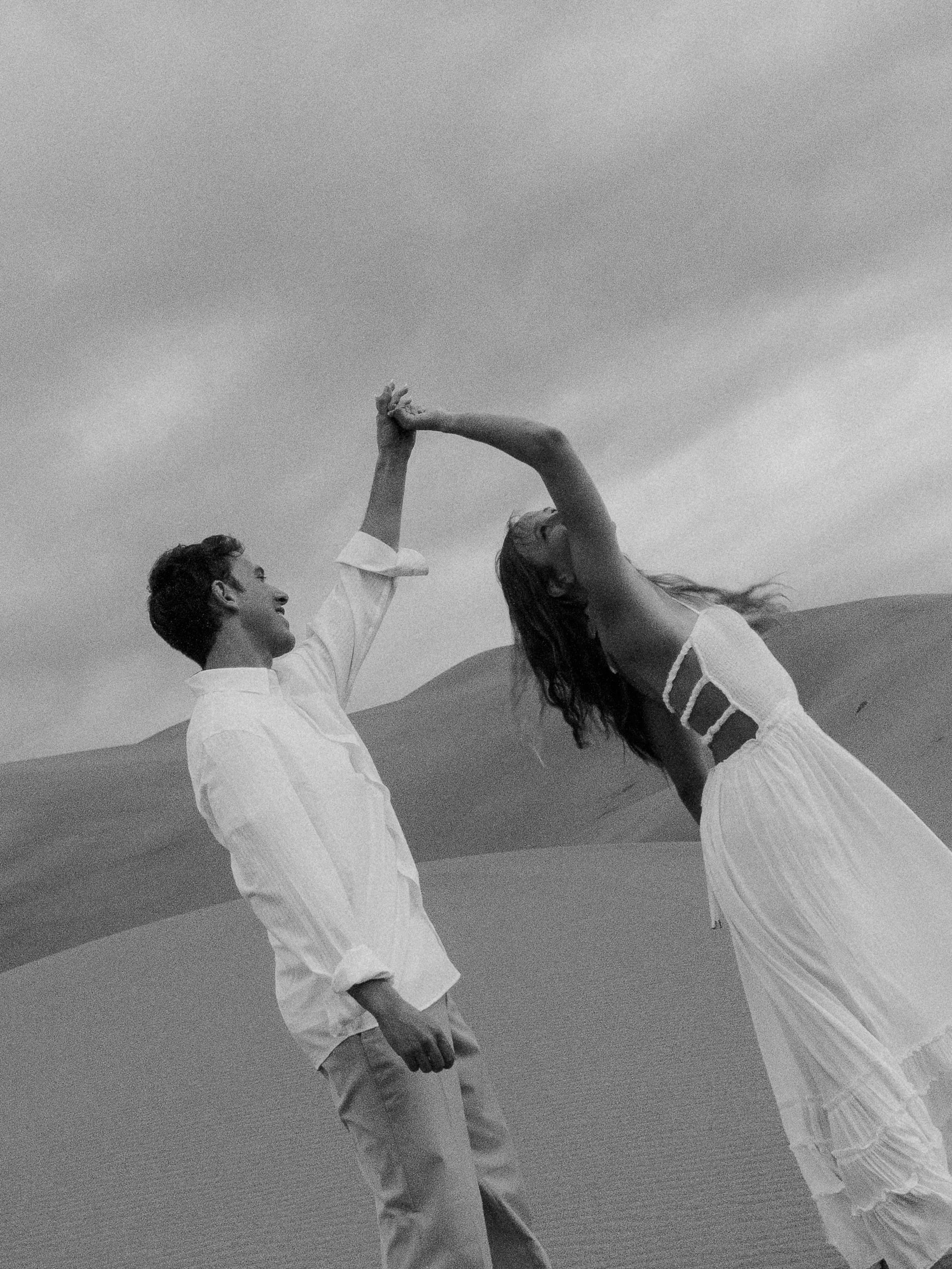 A couple dancing in a desert landscape, holding hands and leaning back with one arm raised, dressed in light clothing.