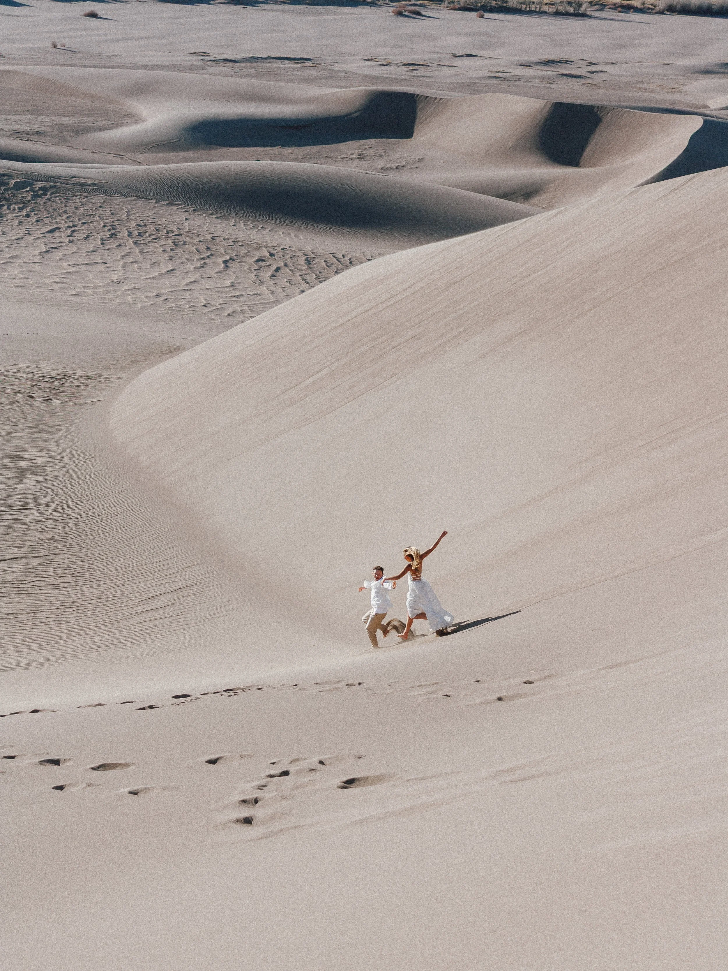 A couple in white clothing running hand-in-hand on sand dunes in a desert landscape.