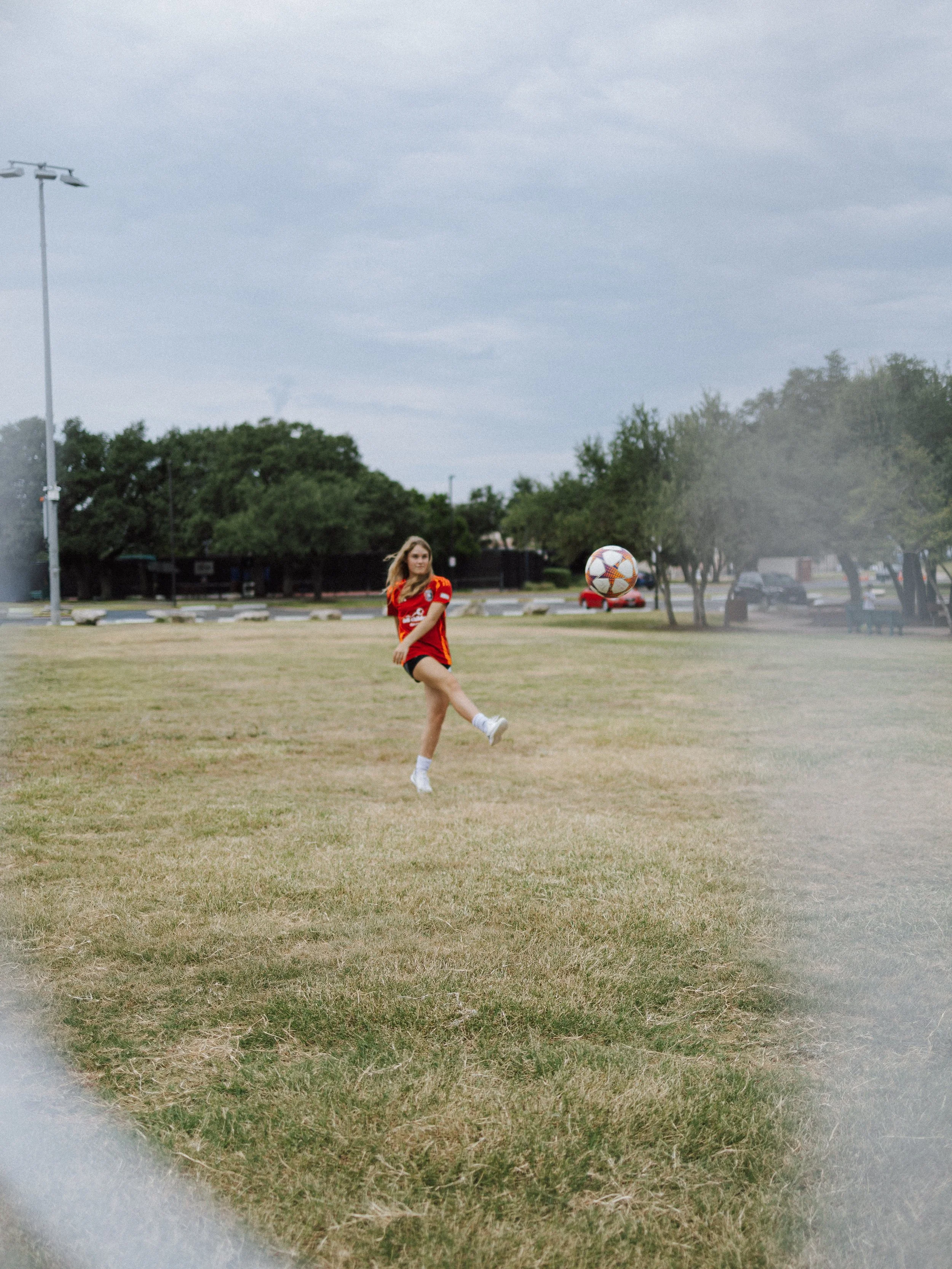 A woman in a red sports jersey and black shorts is playing soccer on a grassy field, with a soccer ball nearby. Behind her are trees, parked cars, and a cloudy sky.
