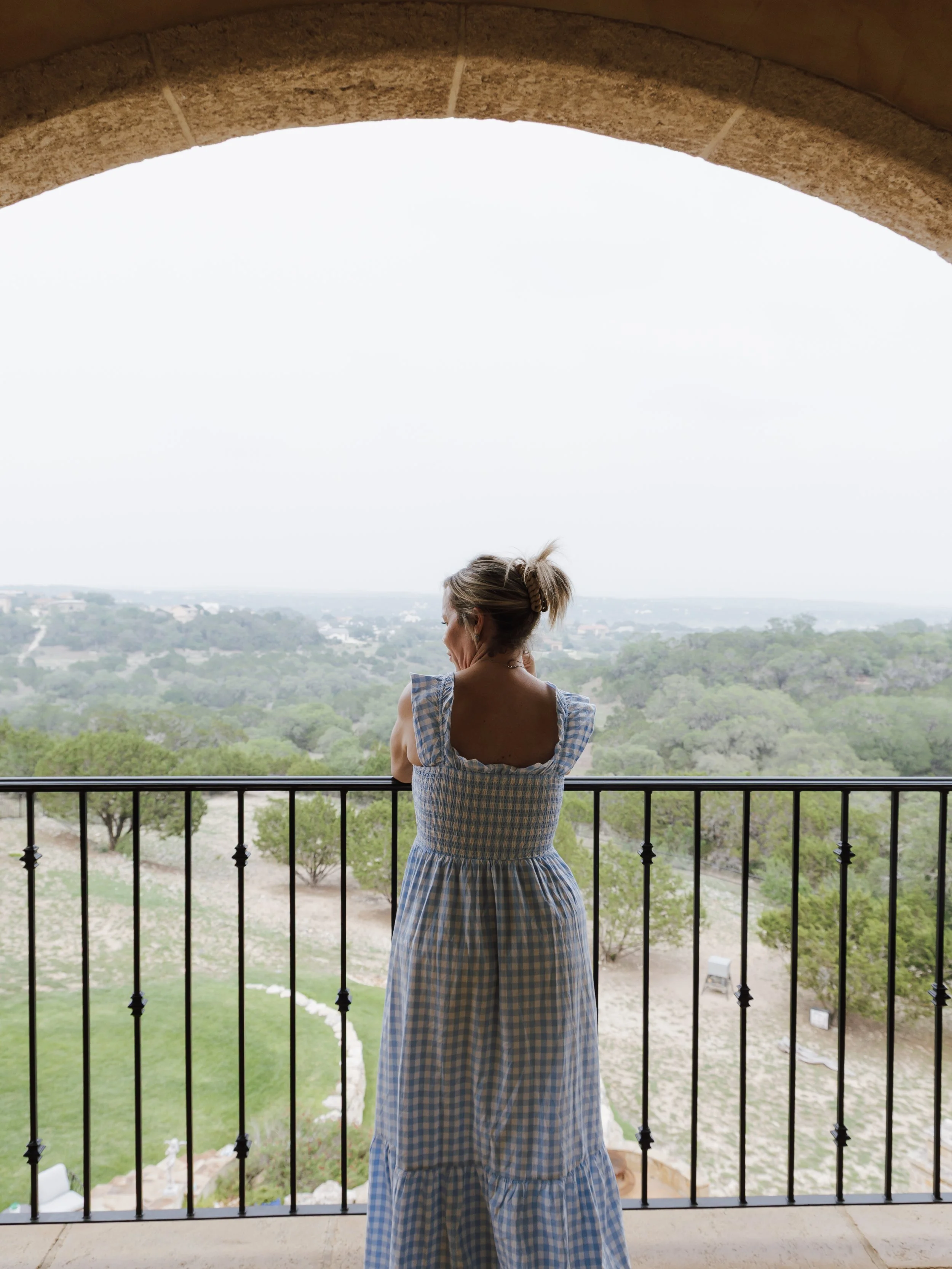 A woman in a blue and white checkered dress standing on a balcony with a view of trees and a cloudy sky.