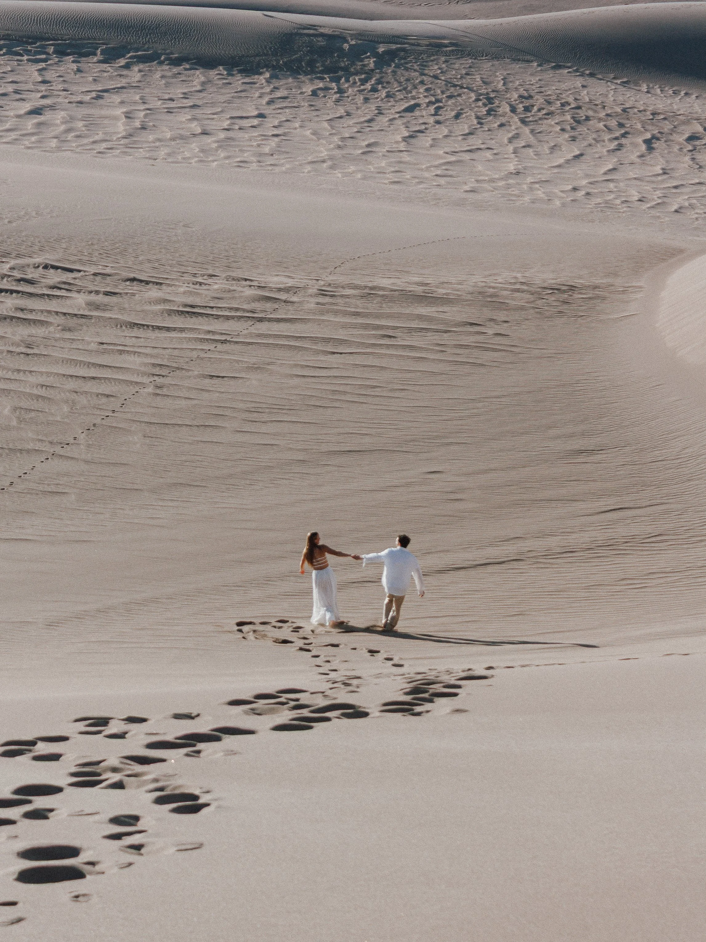 A man and woman walking hand in hand across sand dunes, leaving footprints behind, with a vast desert landscape in the background.