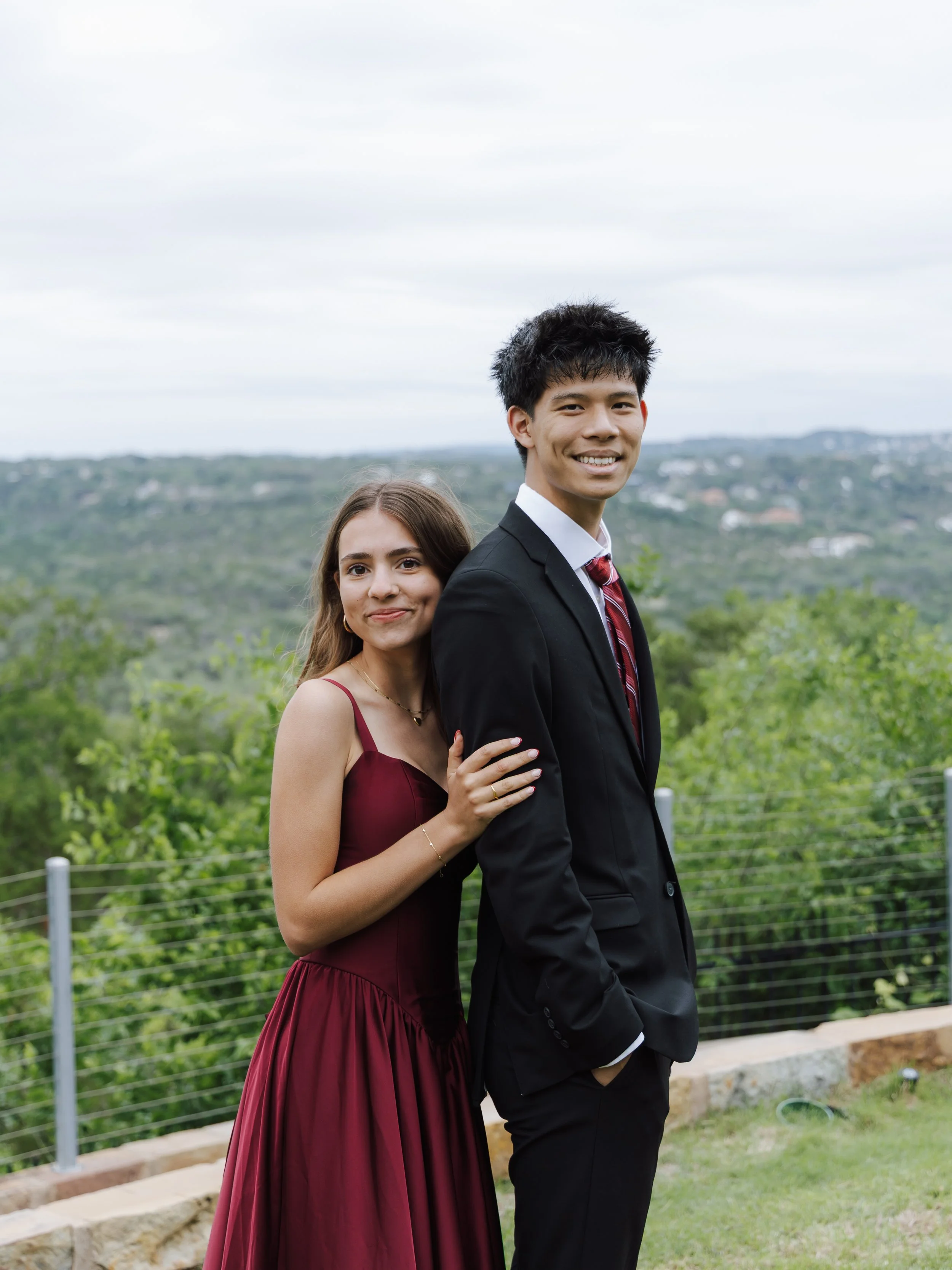 A young woman in a burgundy dress standing next to a young man in a black suit, outdoors with green trees and a cloudy sky in the background.