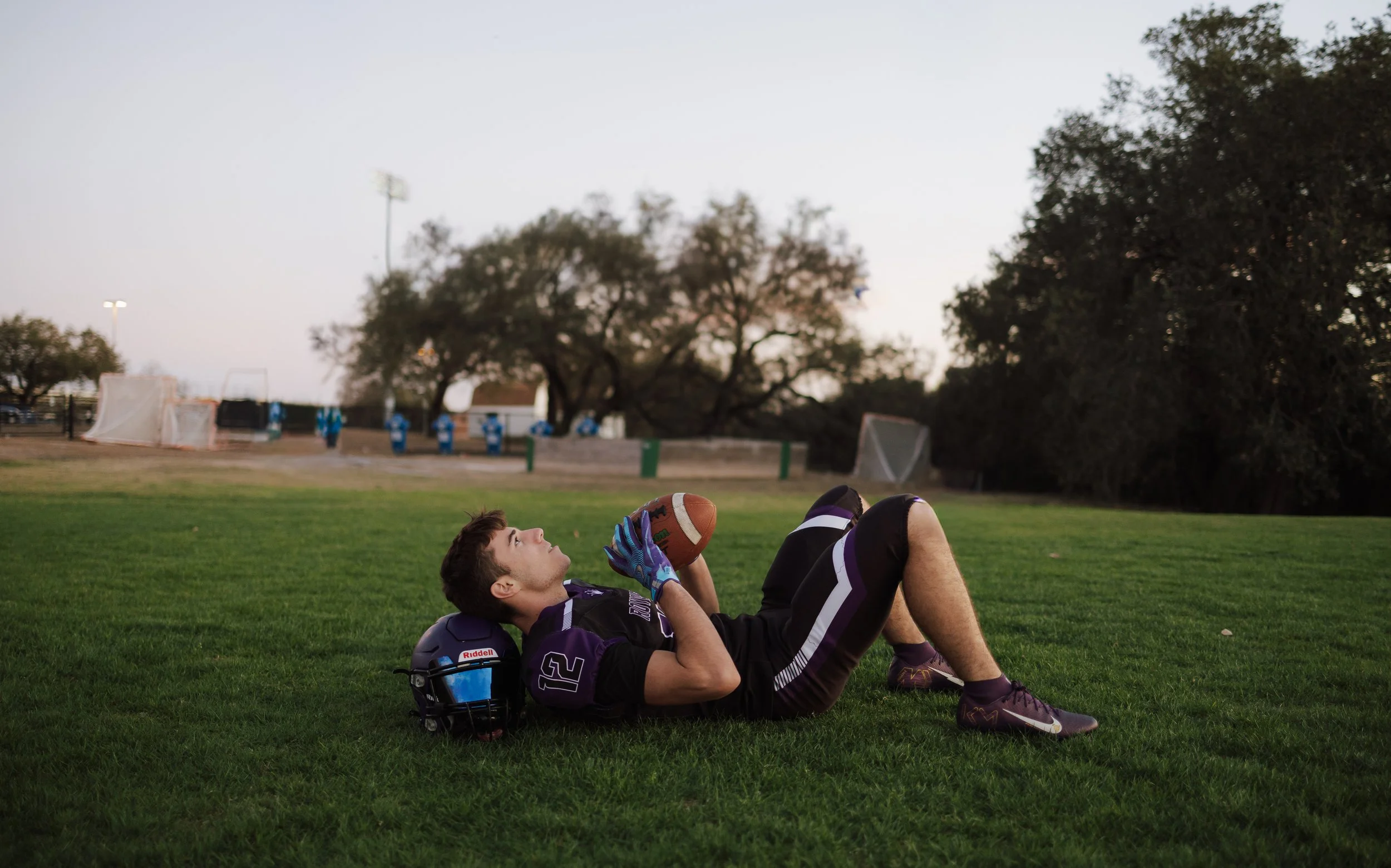 Football player lying on the grass with helmet and gloves beside him, holding a football in his hands, on a sports field during twilight.