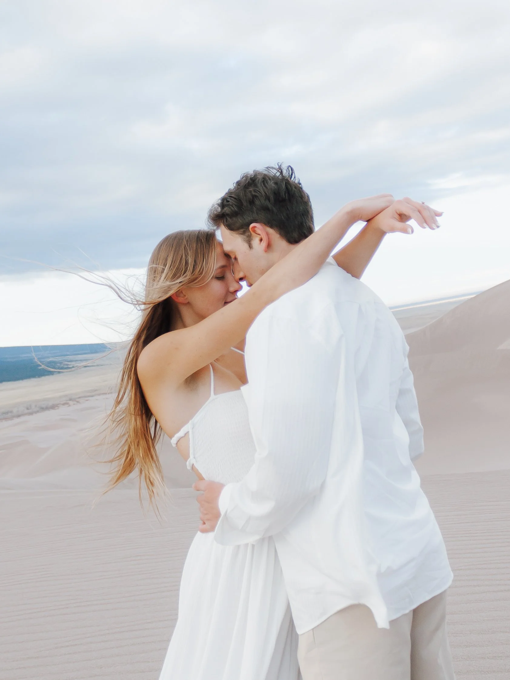 A couple embracing and touching foreheads in a desert with sand dunes under a cloudy sky.