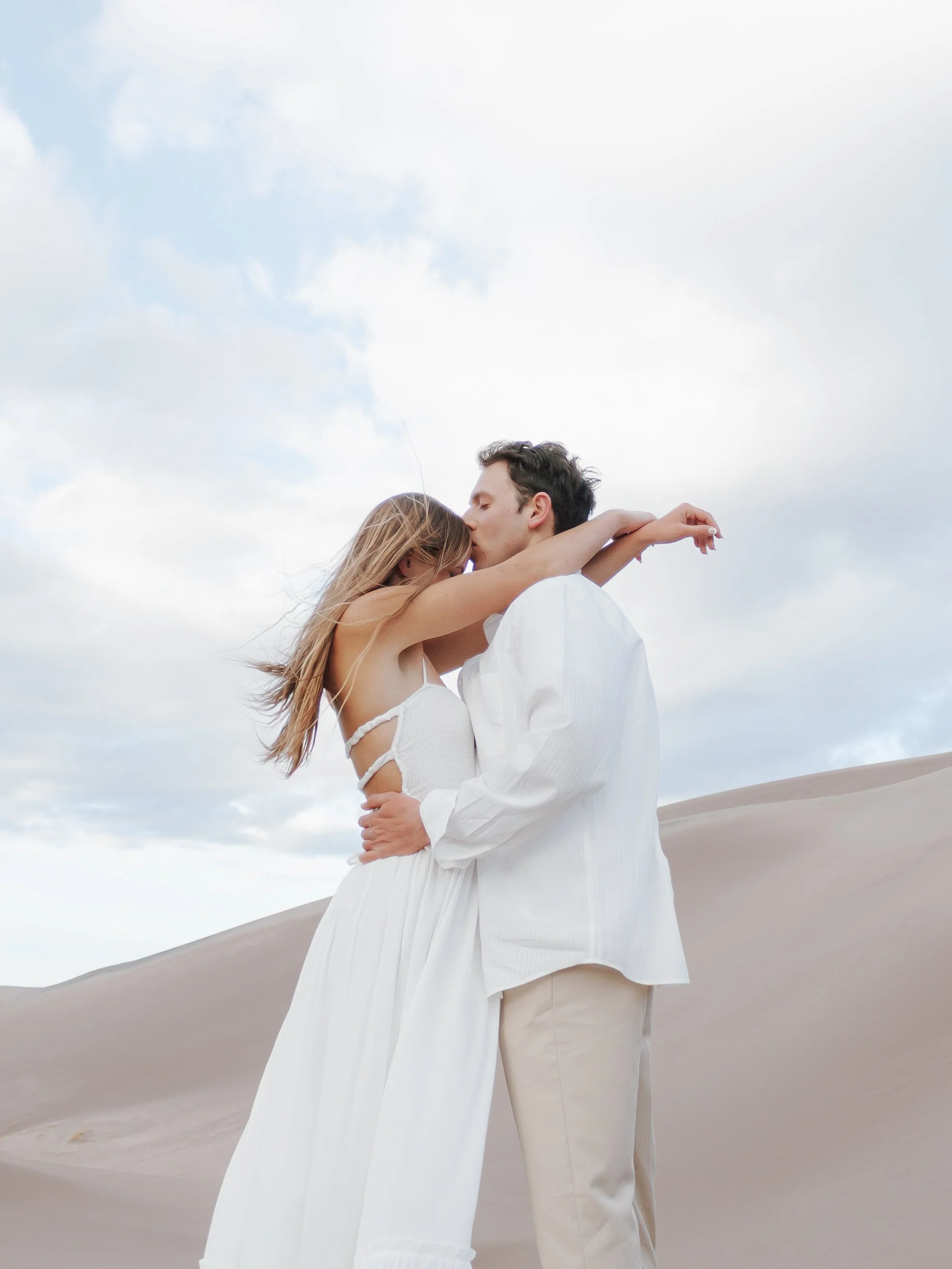 A couple embraces and kisses in a desert landscape with sand dunes and a cloudy sky.